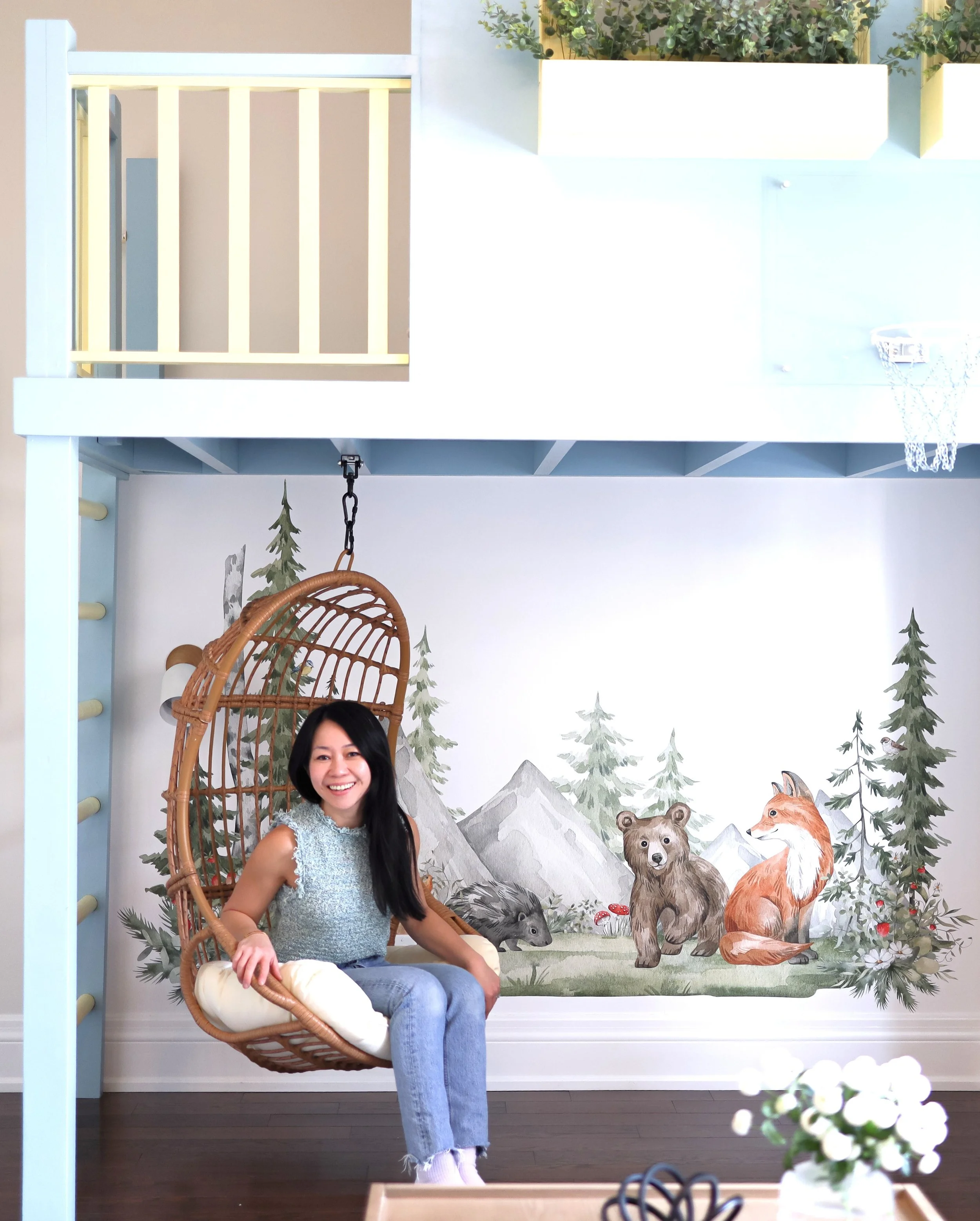 Smiling woman sitting in a hanging wicker chair in front of a woodland theme mural featuring bears, a fox, and pine trees, with a white cushion on her lap and a small bouquet of white flowers in the foreground.