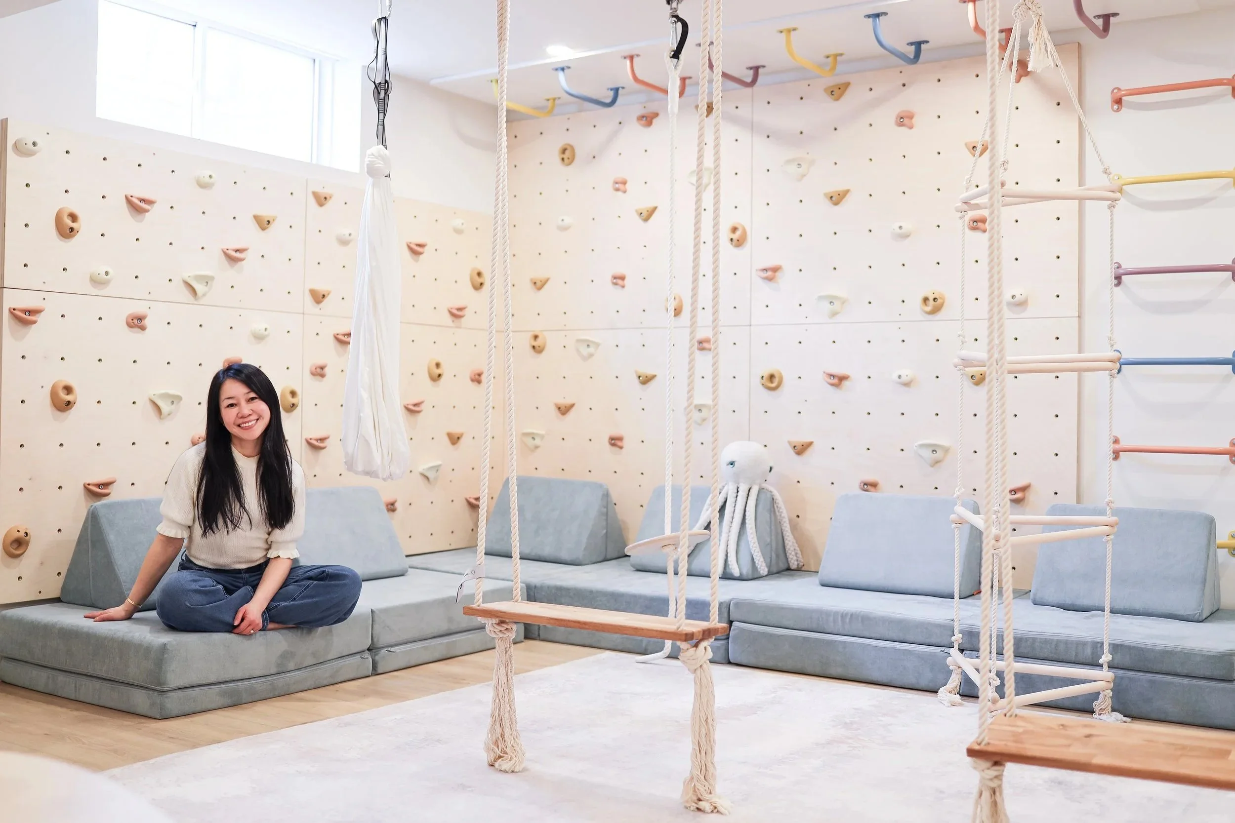 Young woman sitting cross-legged on a gray cushioned floor mat in a room with a climbing wall and indoor swings.