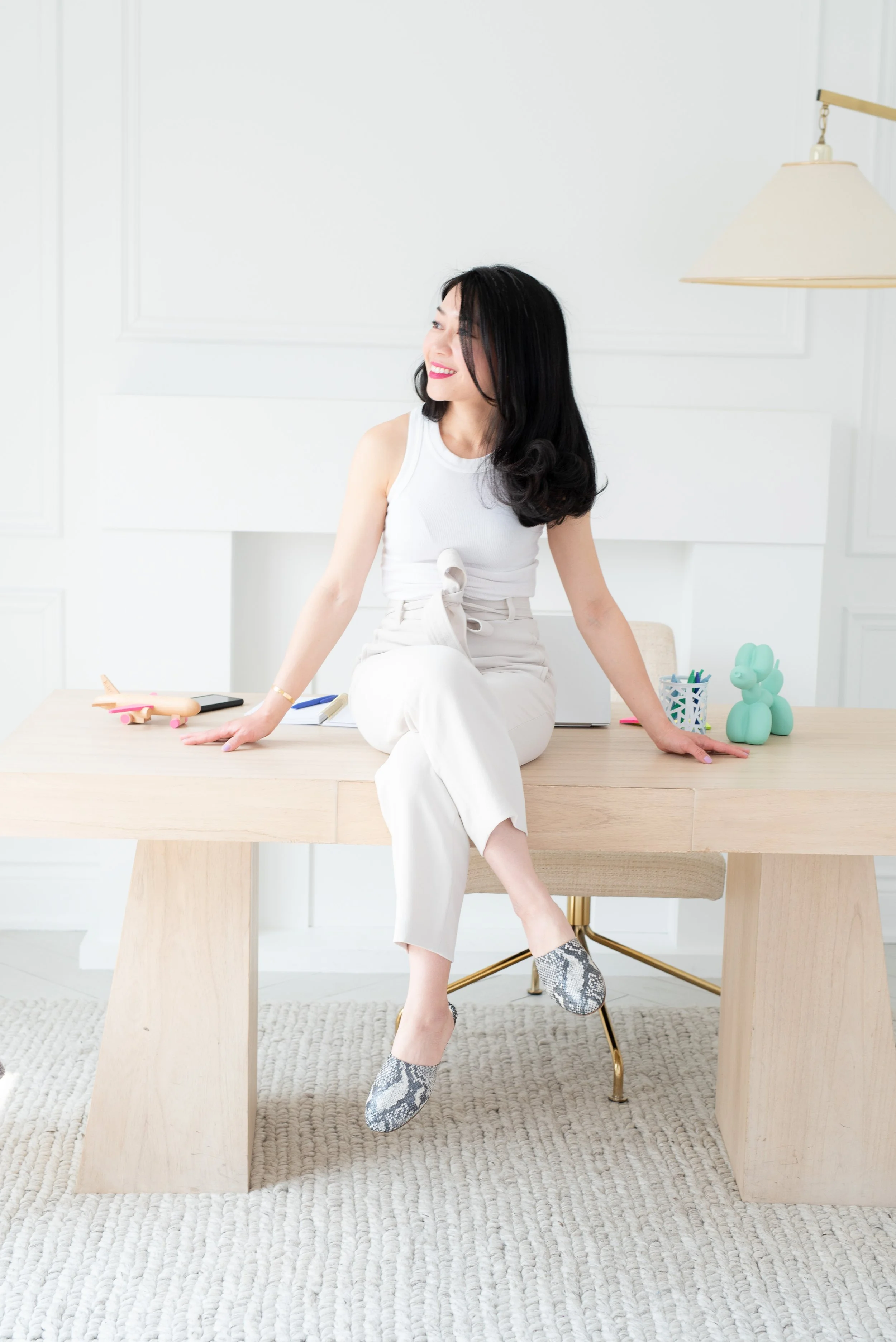 A woman with black hair, sitting on a wooden table, smiling and looking to her left, in a bright white room with a lamp hanging from the ceiling, a turquoise bunny sculpture, and stationery on the table.