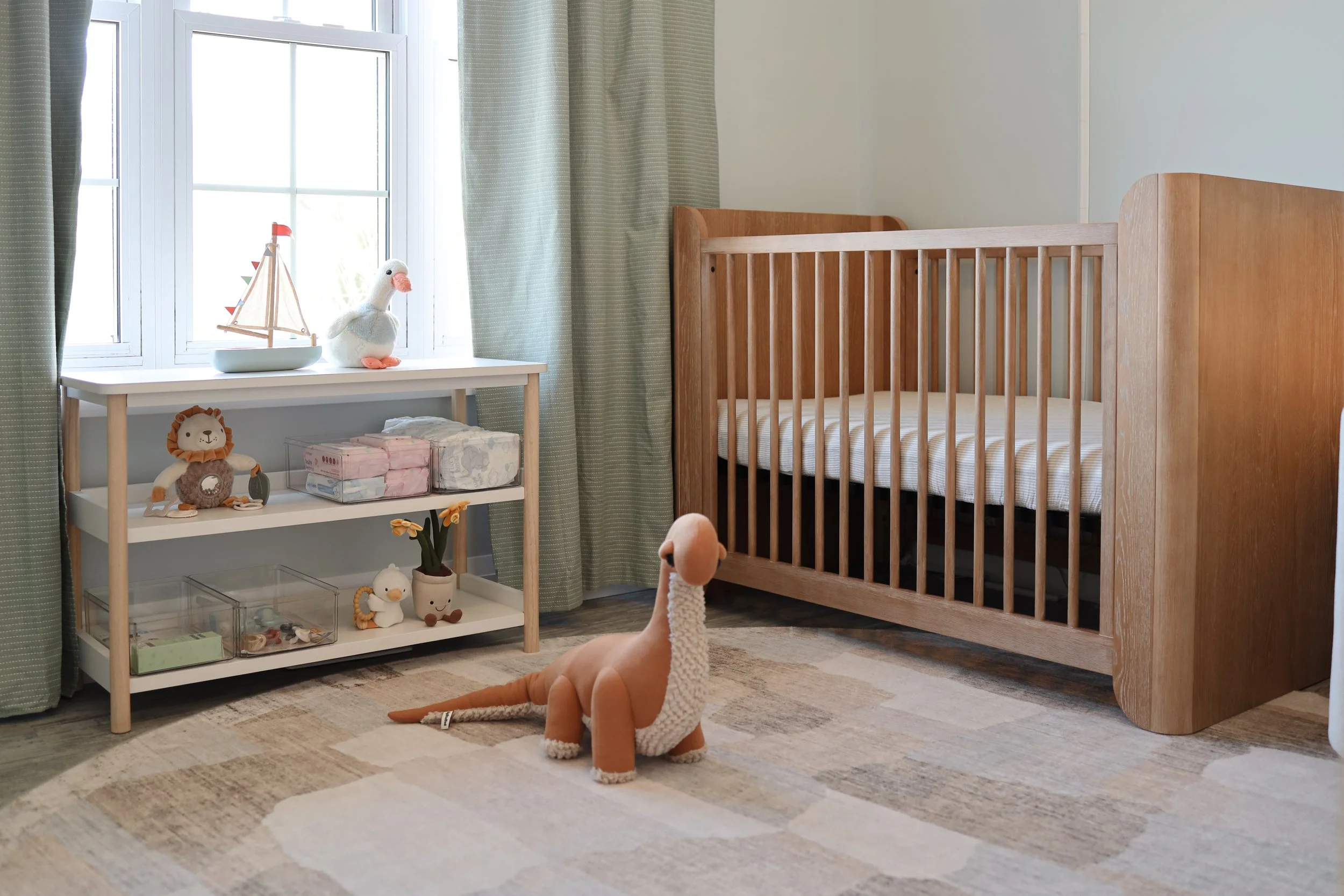 A nursery with a wooden crib, a plush dinosaur toy on a beige patterned rug, a white shelf with stuffed animals, toys, tissue box, and a decorative plant, and a window with curtains.