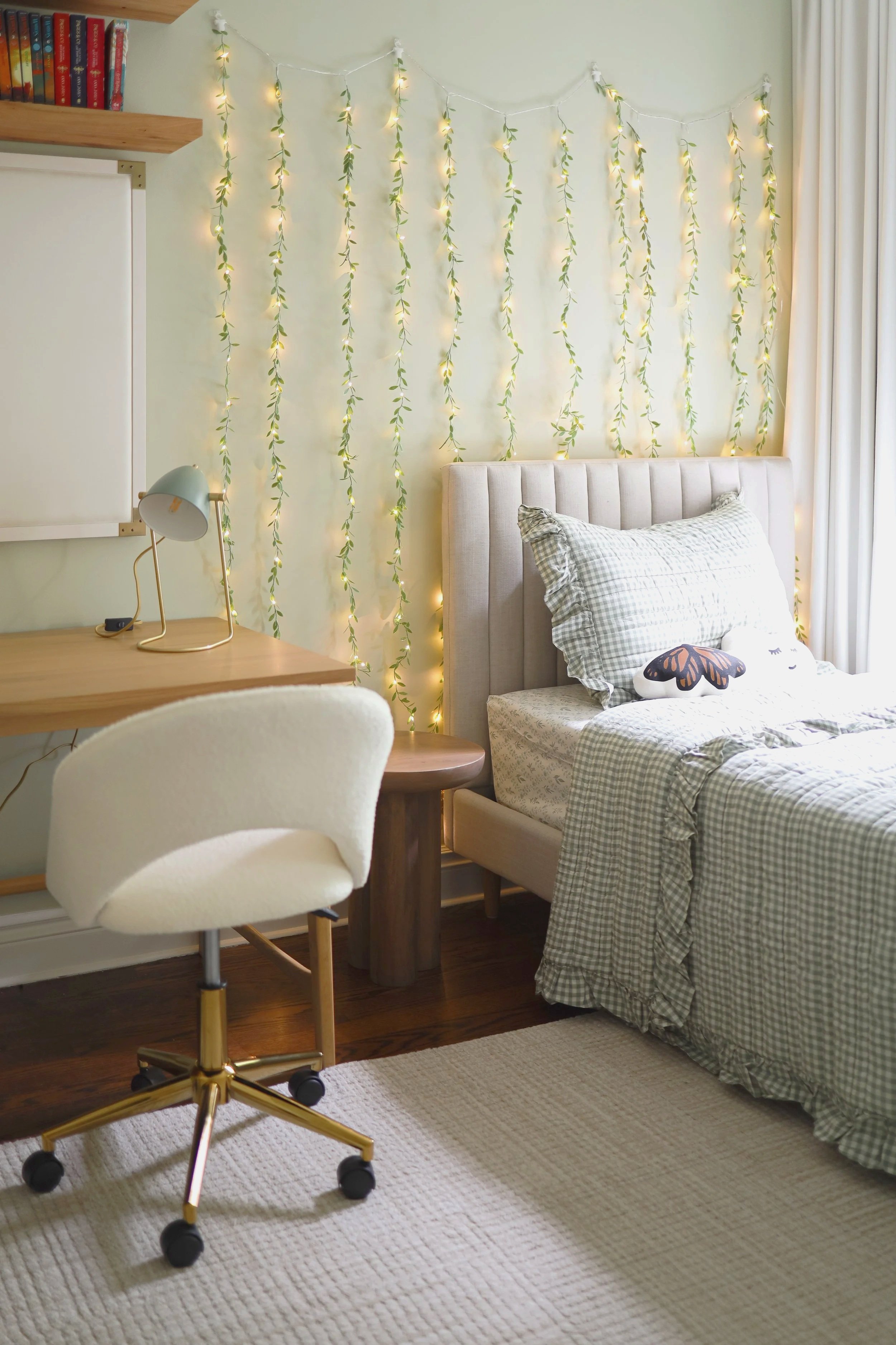 A cozy bedroom corner with a bed, pillows, a string of fairy lights with leaves on the wall, a desk, a white chair with gold legs, a table lamp, a bookshelf with books, and a window with light curtains.