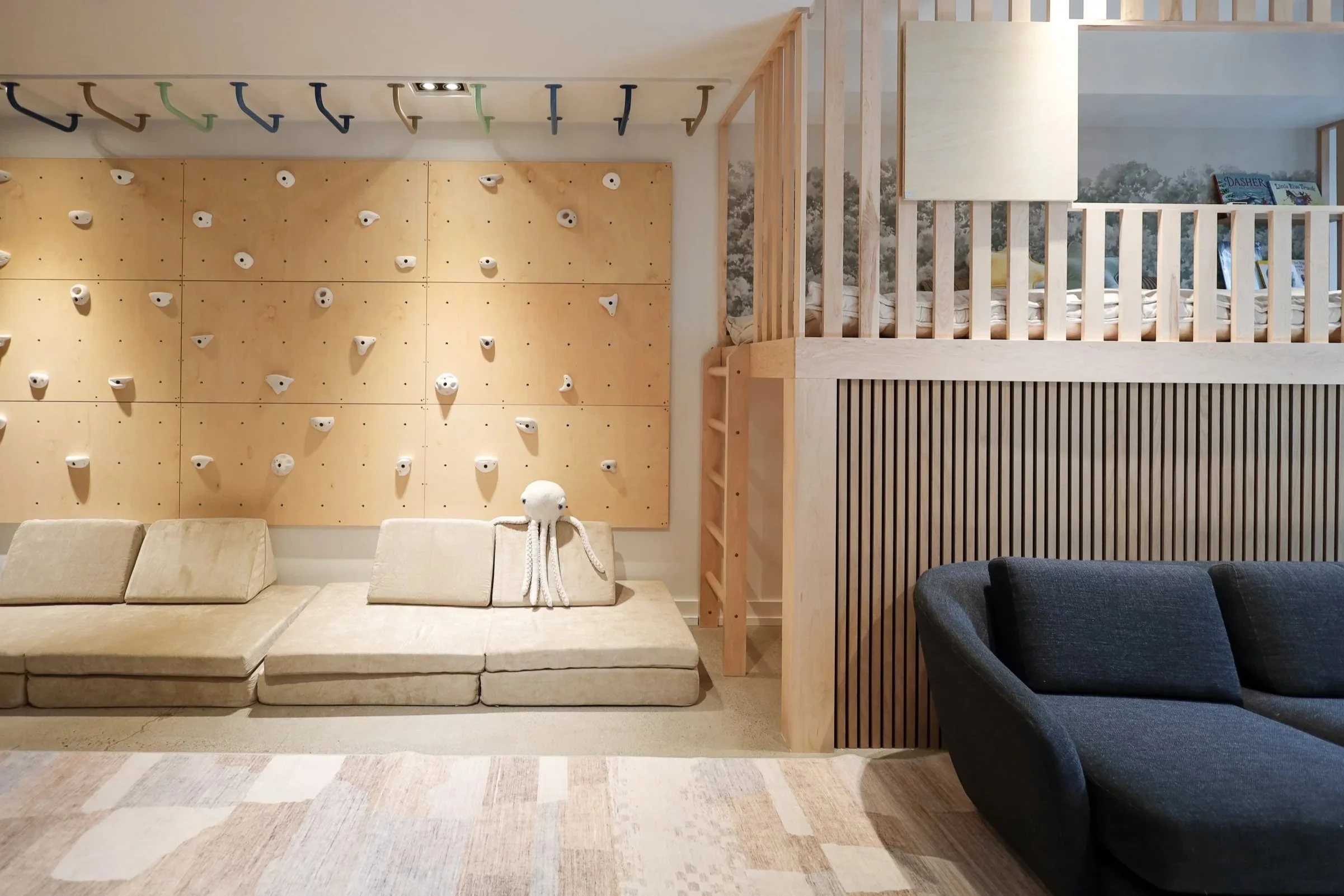 Living room with beige sectional sofa, climbing wall on yellow wooden panel, and partially visible loft area with books and mural of trees.