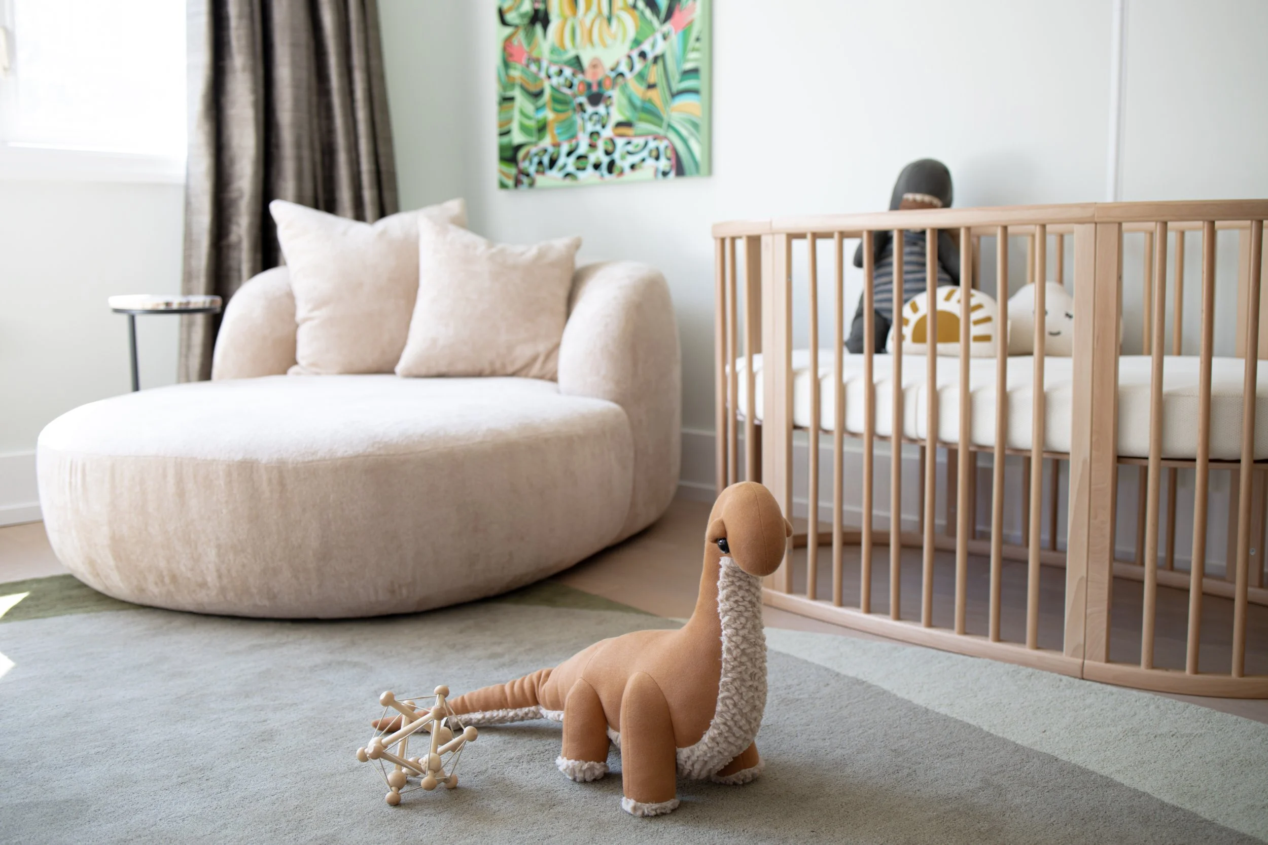 A nursery with a wooden crib, a plush dinosaur toy on a beige patterned rug, a white shelf with stuffed animals, toys, tissue box, and a decorative plant, and a window with curtains.
