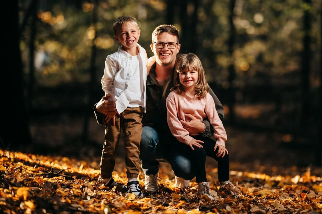 Dad kneeling with his son and daughter on golden fallen leaves in backlit woods, Concord NH family portraits