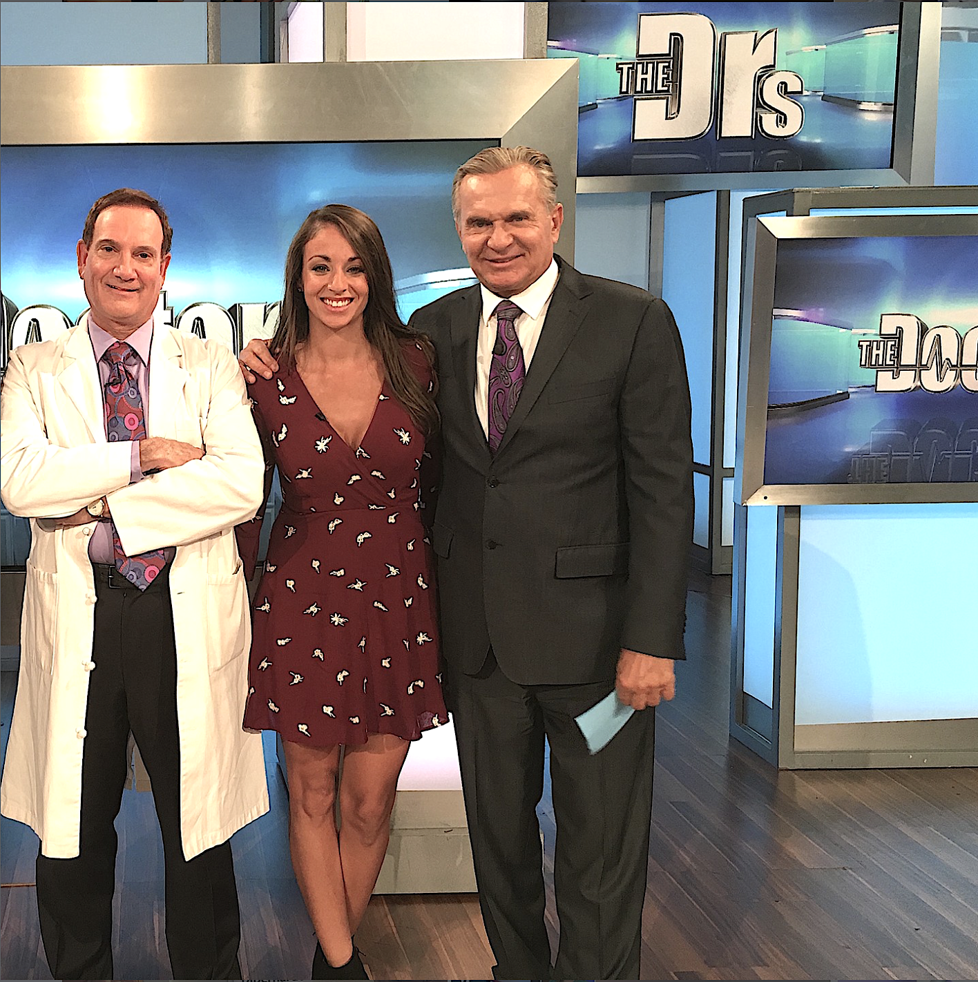 Dr. Barry DiBernardo with "The Doctors" show hosts standing together onstage, smiling for the camera. The background features a large sign reading 'The Drs' and studio monitors.