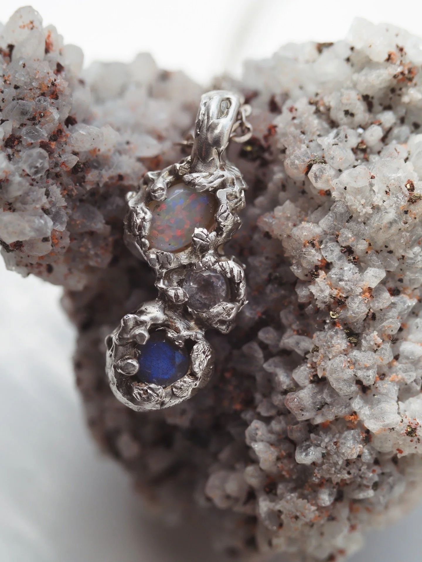 A silver ring with embedded opal and blue gemstone set in a textured silver band, resting on coarse white and gray gravel.