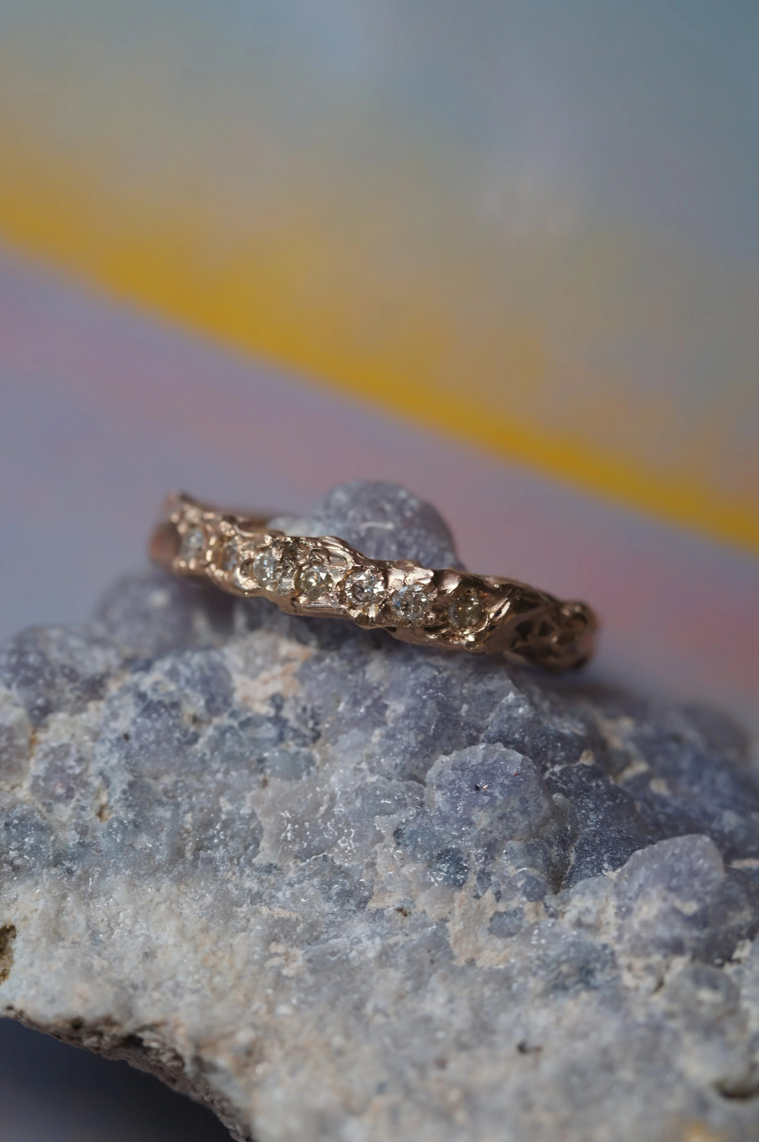 A close-up of a gold ring with embedded diamonds, placed on a rock surface with a blurred background of pastel yellow, pink, blue, and gray hues.