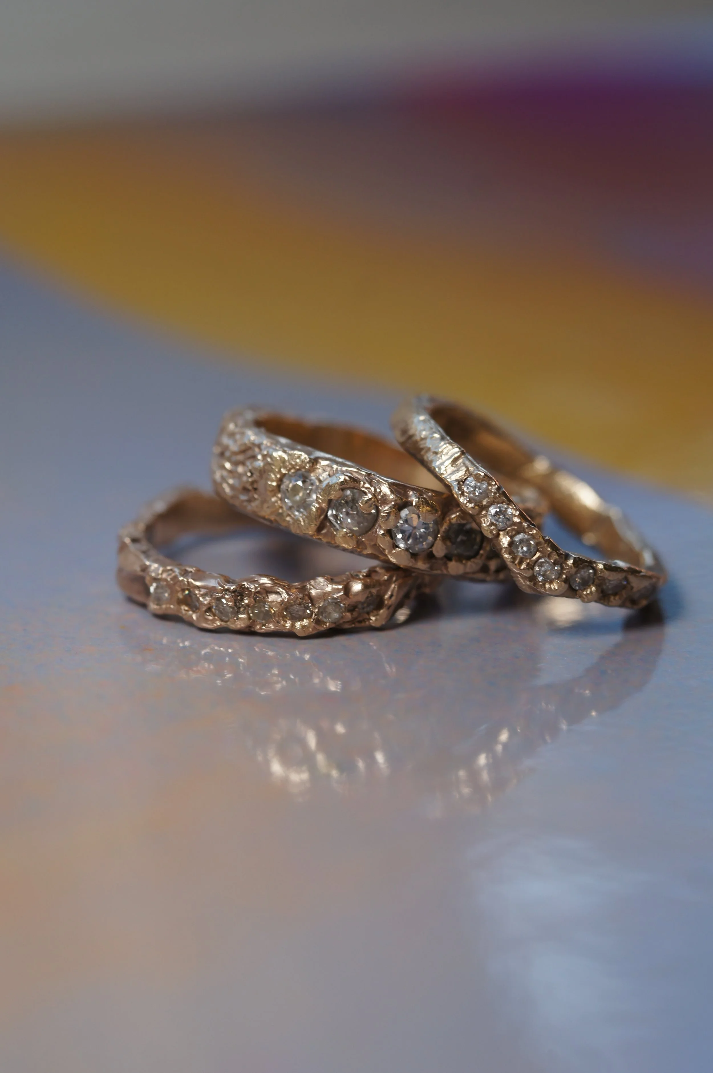 Close-up of three intertwined gold rings adorned with small diamonds, on a reflective surface with a blurred, multi-colored background.