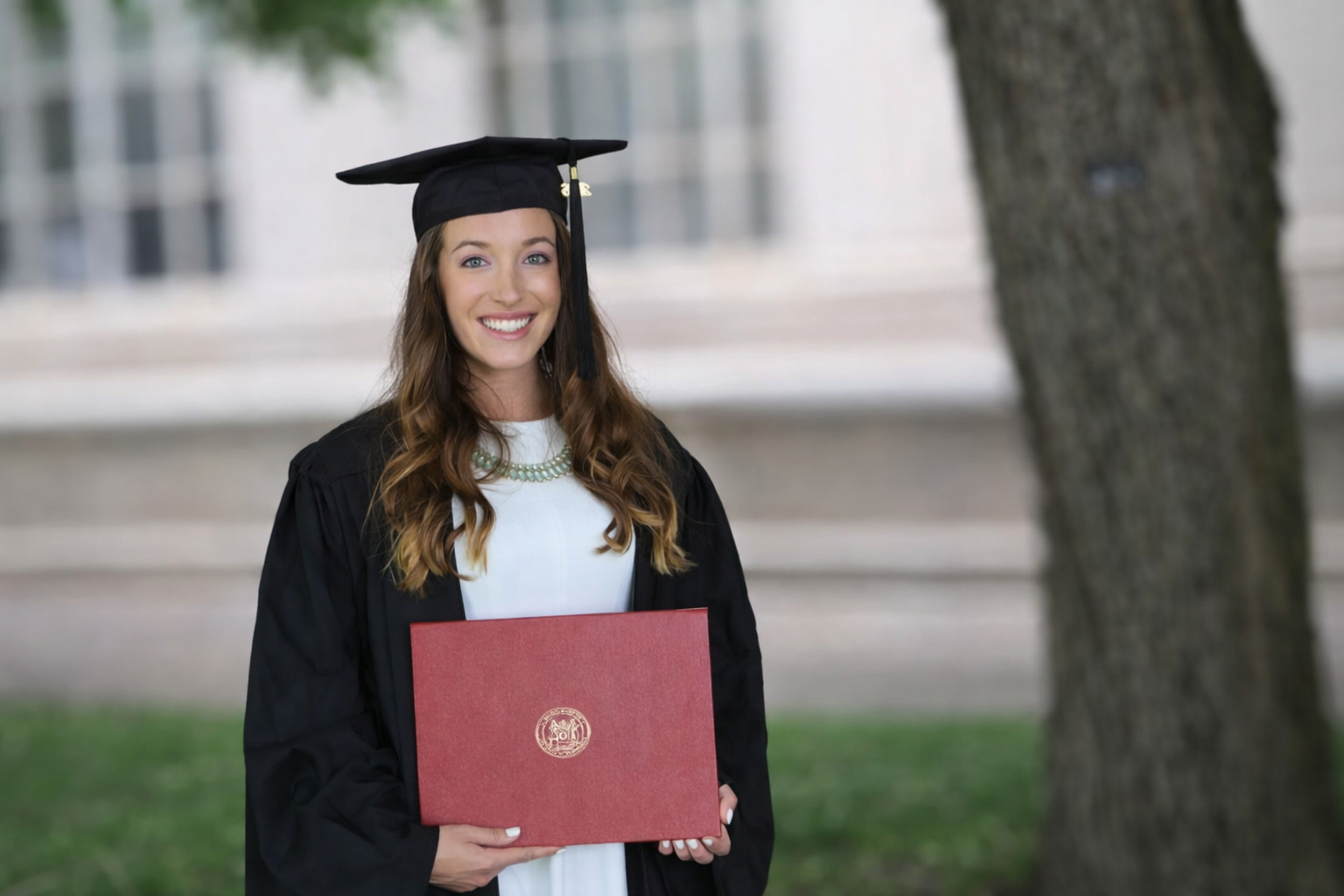 A young woman in a graduation cap and gown smiling and holding a diploma outside near a building with large windows.