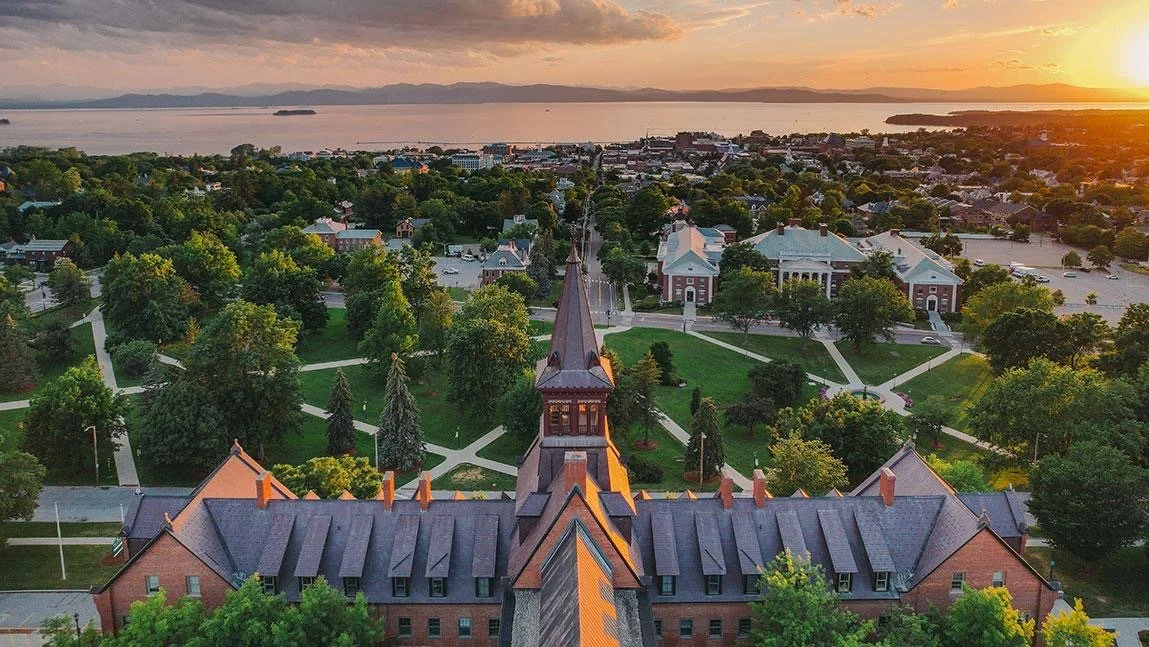 Aerial view of a college campus at sunset with a prominent brick building and a tall steeple in the foreground, surrounded by green trees, paths, and additional buildings near a body of water.