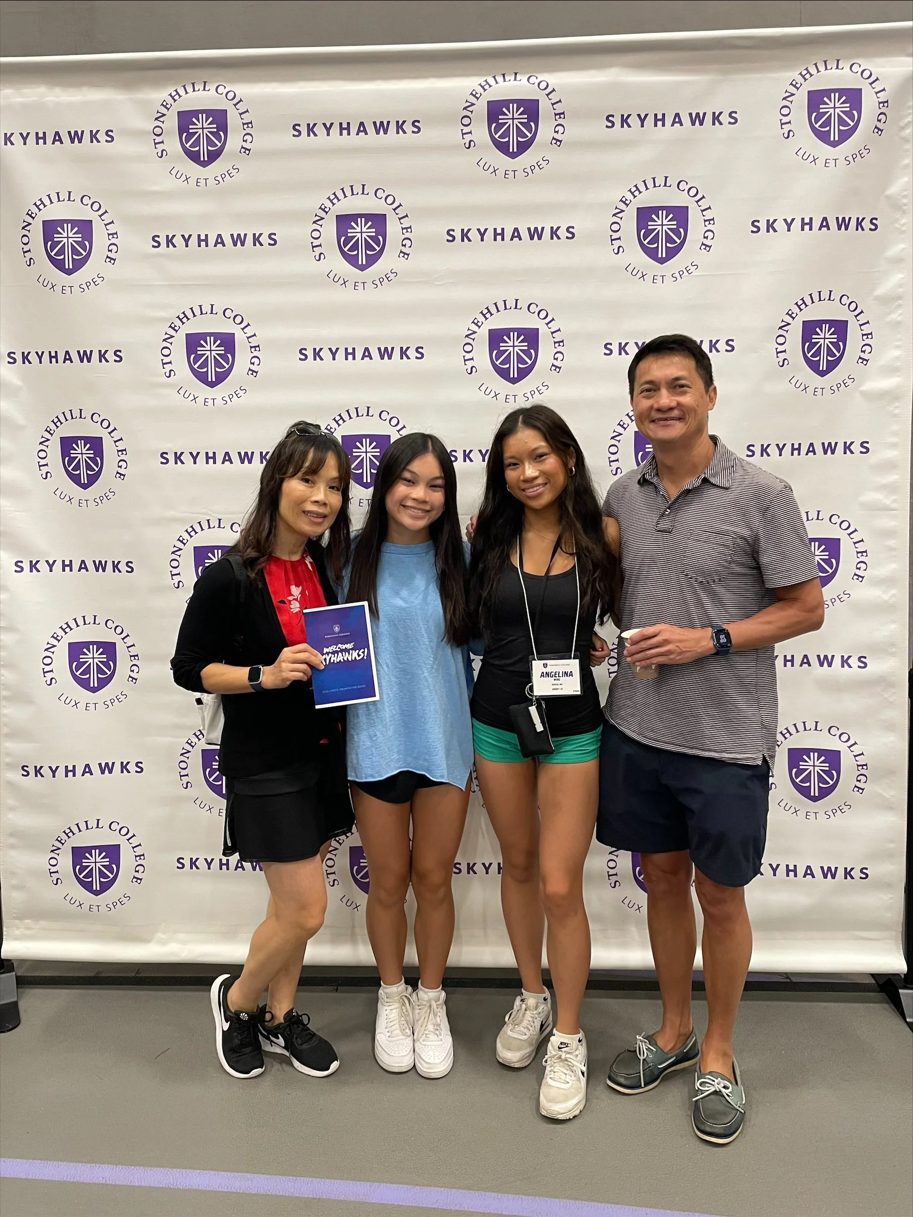 Group of four people standing together in front of a Stonehill College Skyhawks backdrop, smiling at the camera. One woman is holding a welcome sign, and another woman has a name tag that reads Angelina.