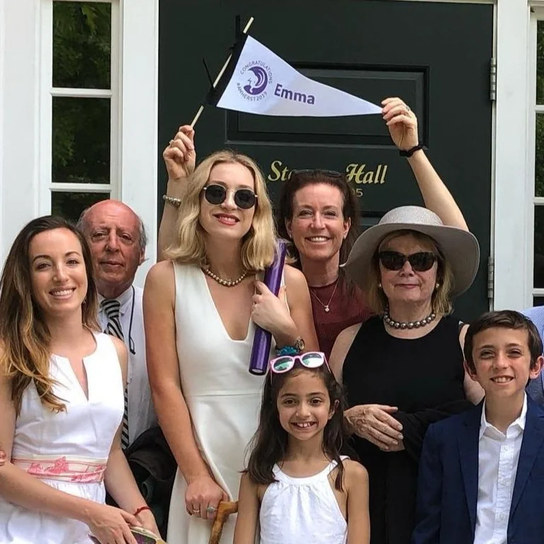 Group of celebrating family members standing outside, with some wearing party accessories and holding a congratulatory flag for Emma, in front of a house with a black door.