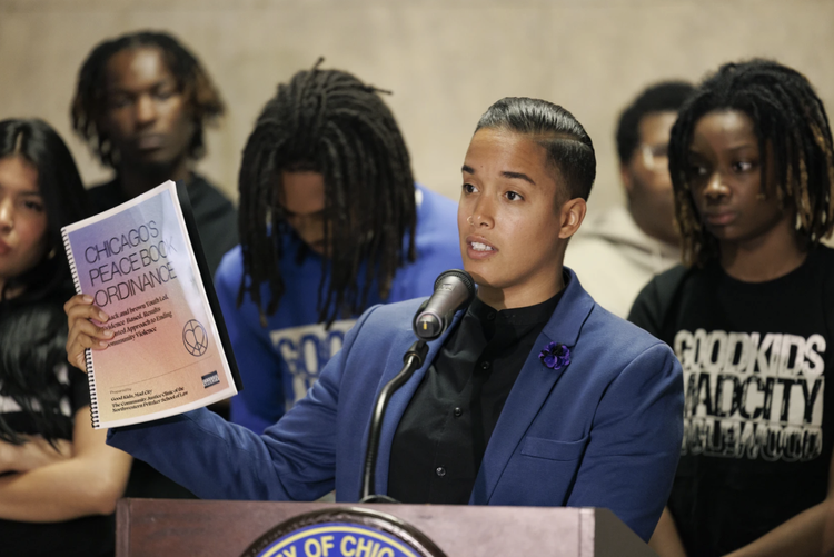 A woman in a blue blazer speaking at a microphone during a public meeting, holding a document titled 'Chagos Peace Book Ordinance,' with a group of people standing behind her.