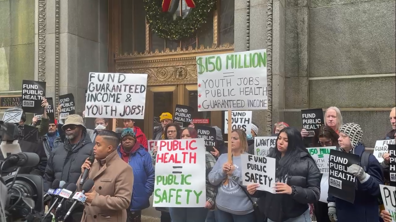 Group of people protesting outside a government building, holding signs demanding public health funding, youth jobs, and guaranteed income, with some signs saying '$150 MILLION', 'FUND PUBLIC HEALTH', 'FUND GUARANTEED INCOME & YOUTH JOBS!', 'PUBLIC HEALTH = PUBLIC SAFETY'.