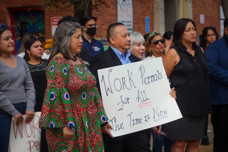 A group of people participating in a protest or rally, with one woman holding a sign that reads 'Work Permits for All The Time is Now'.