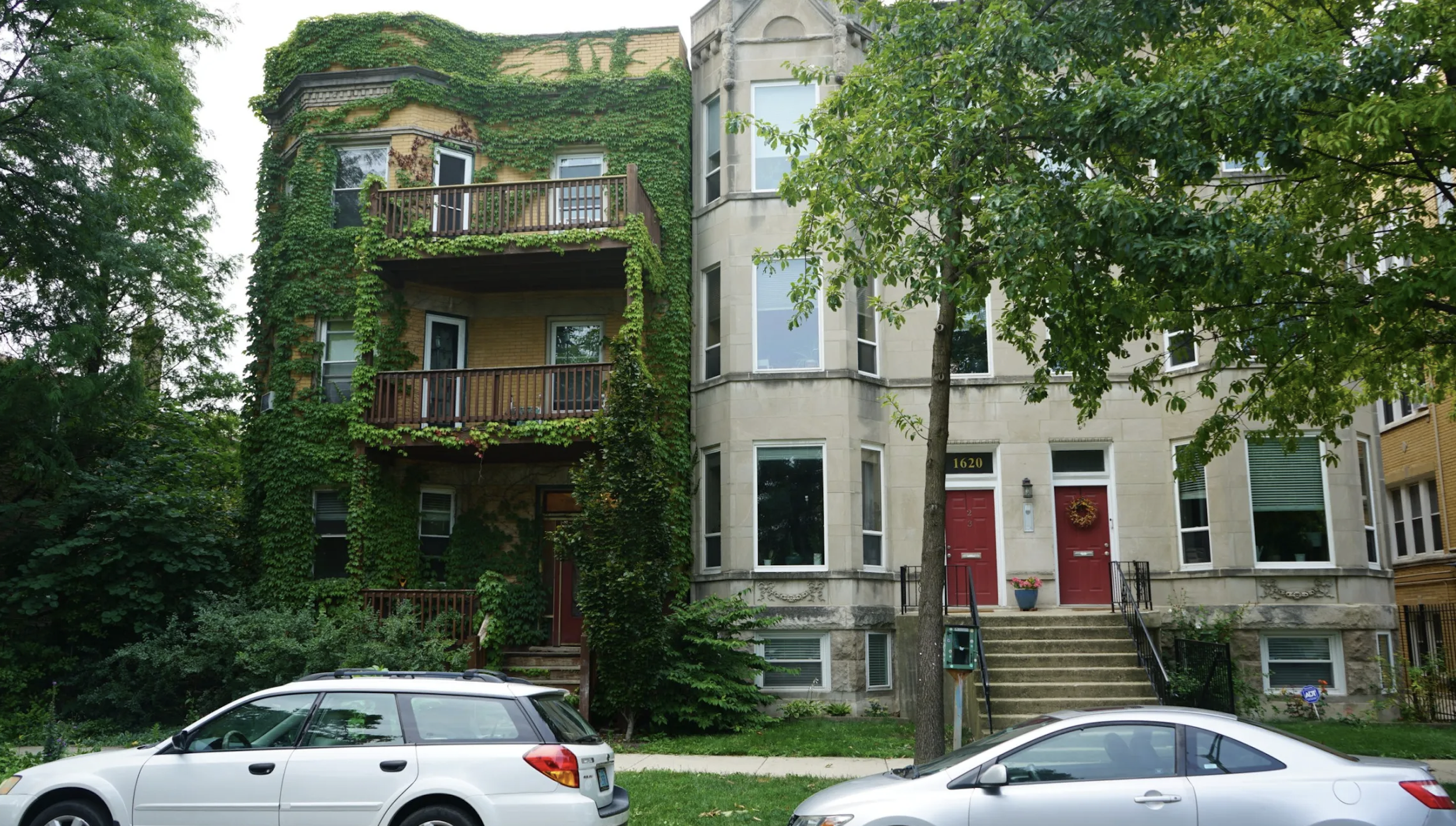 Multi-story residential building with a beige brick facade covered in green ivy, balconies with wooden railings, and a red door with a decorative wreath, situated on a street with parked cars and trees.