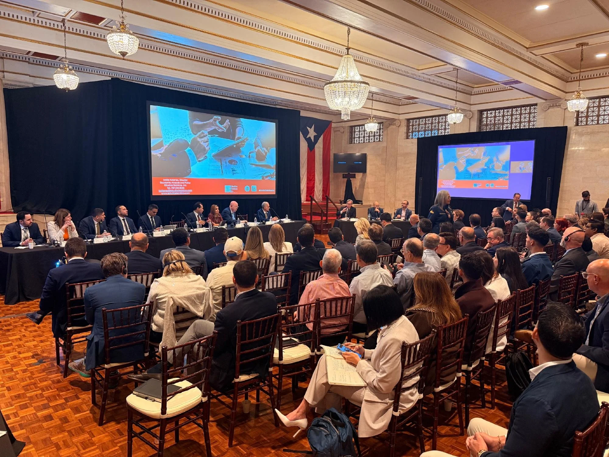 A large conference room filled with people attending a formal event. There are two big screens displaying a presentation, with one showing a close-up of hands using a digital tablet. Attendees are seated at tables, listening, taking notes, or looking at their phones. The room has chandeliers and a Puerto Rican flag in the background, with ornate walls and a wooden floor.