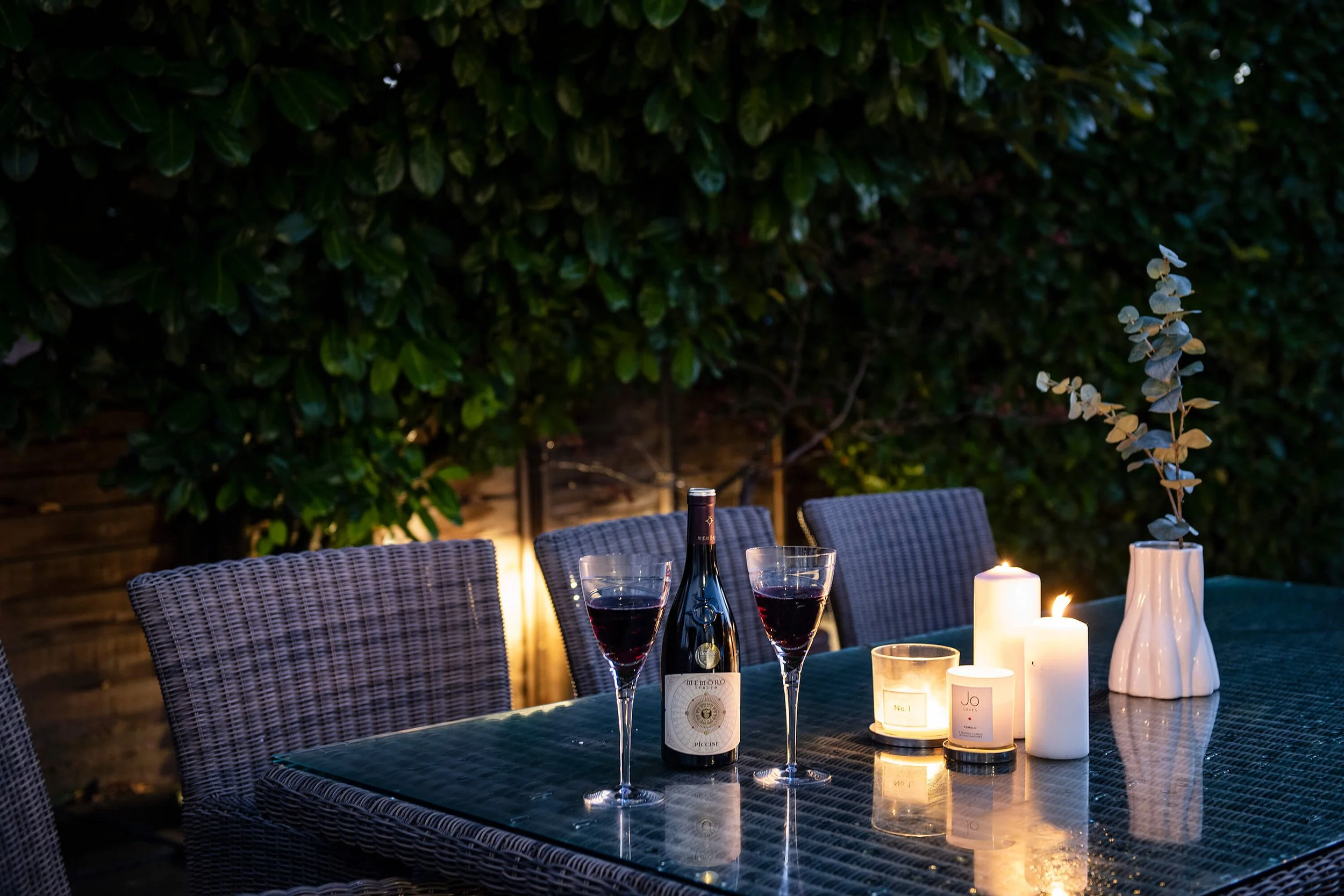 An outdoor dining table with two glasses of red wine, a bottle of wine, and lit candles in a cozy backyard setting at night.