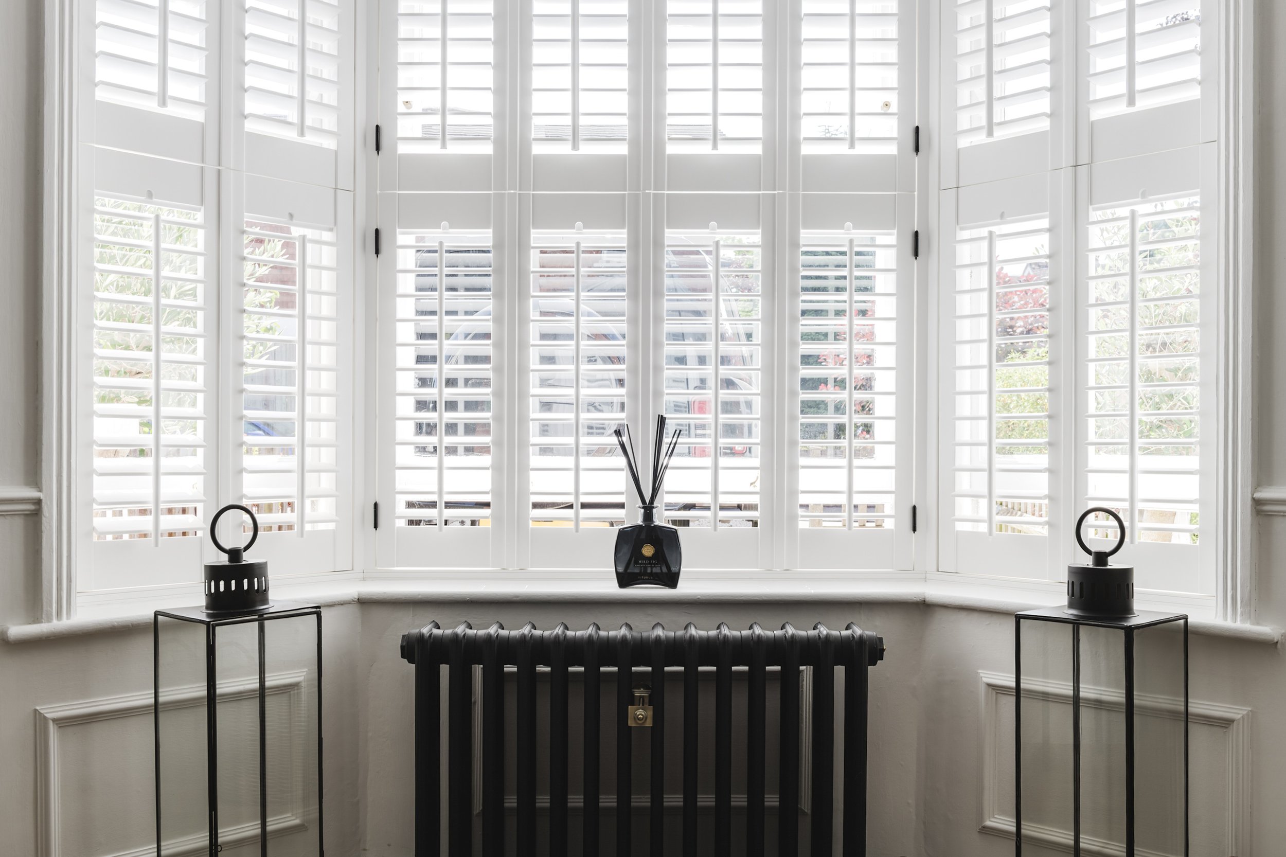 Dining area with a white table and four dark gray chairs, set with plates, cups, and a flower arrangement. There is a cabinet with a plant on top, a wall poster, and an open sliding door leading to a garden with outdoor furniture.