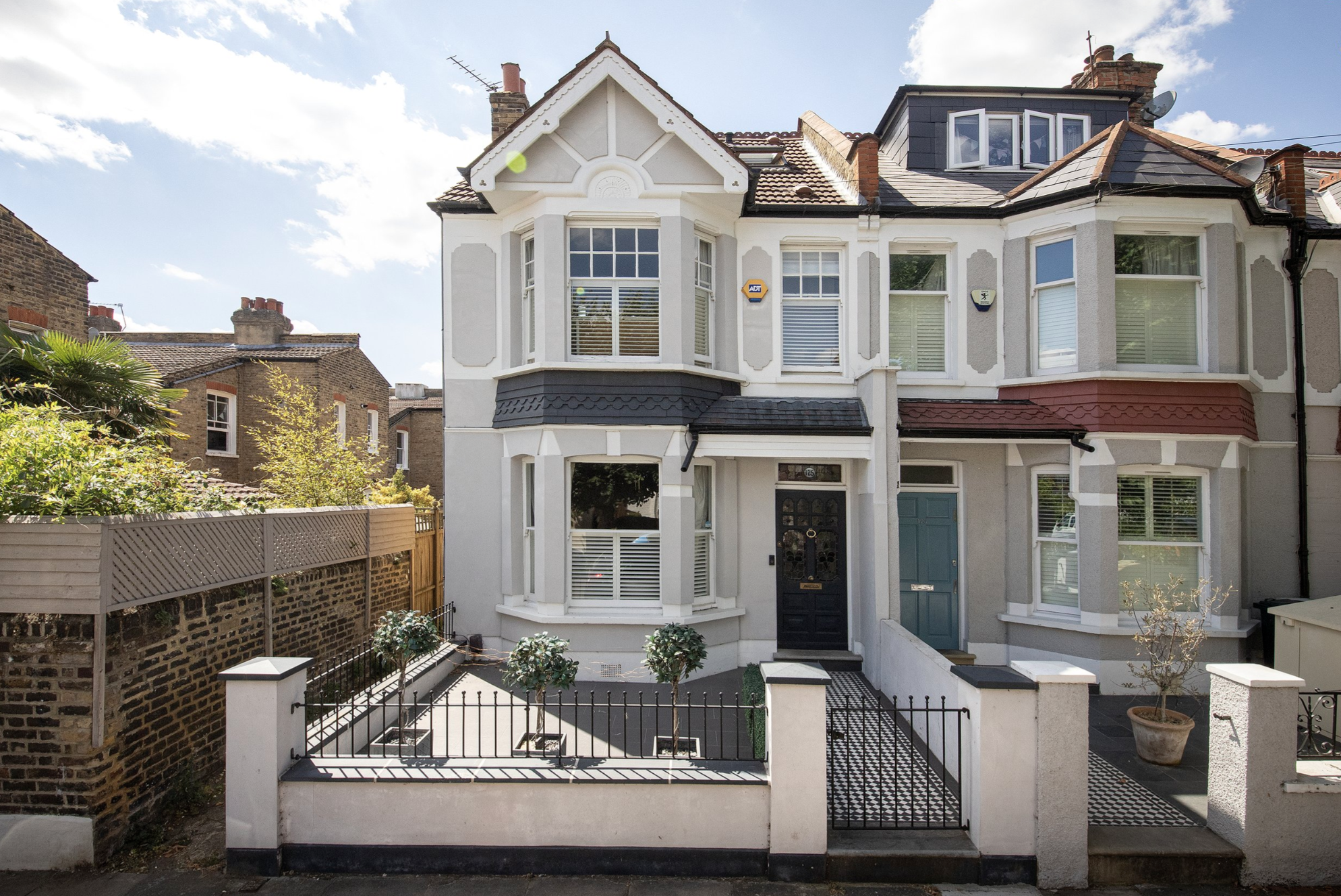 A multi-story Victorian-style house with white exterior walls, bay windows, a black front door, and a small front yard enclosed by a low white wall and black metal fence.