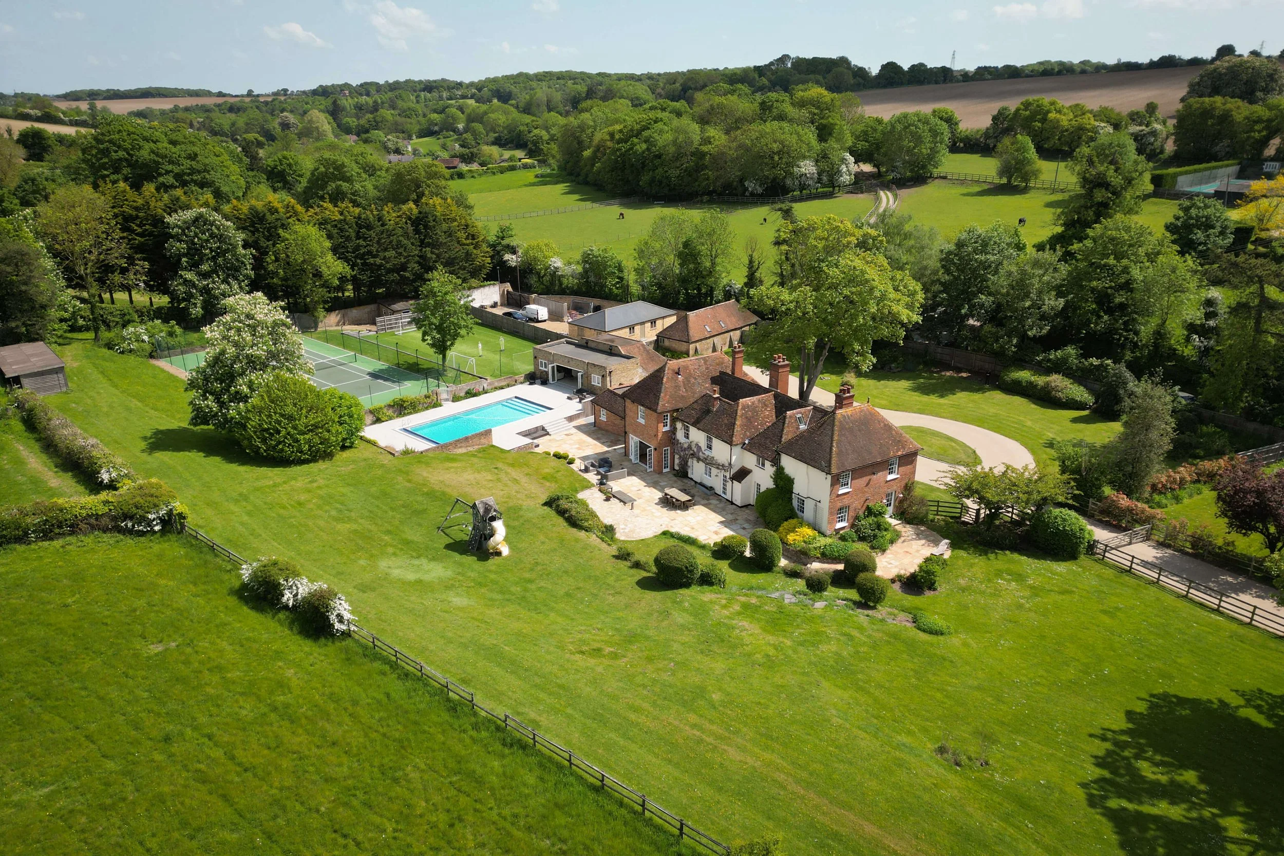 Aerial view of a large countryside estate with a house, swimming pool, tennis court, expansive lawn, trees, and surrounding farmland.