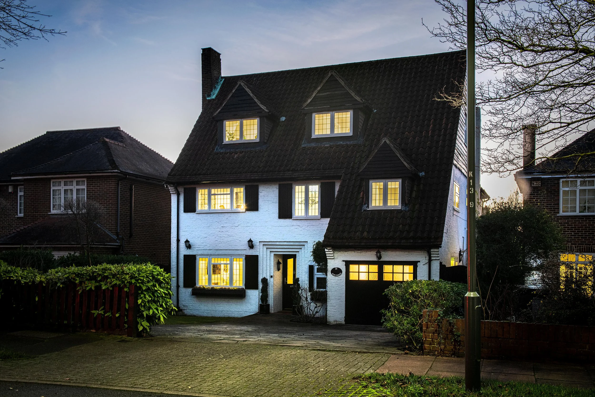 A house with a black tiled steep roof and white walls, illuminated from inside, at dusk. Windows have black shutters, and the house has a brick driveway and a small garden with shrubbery and a wooden fence.