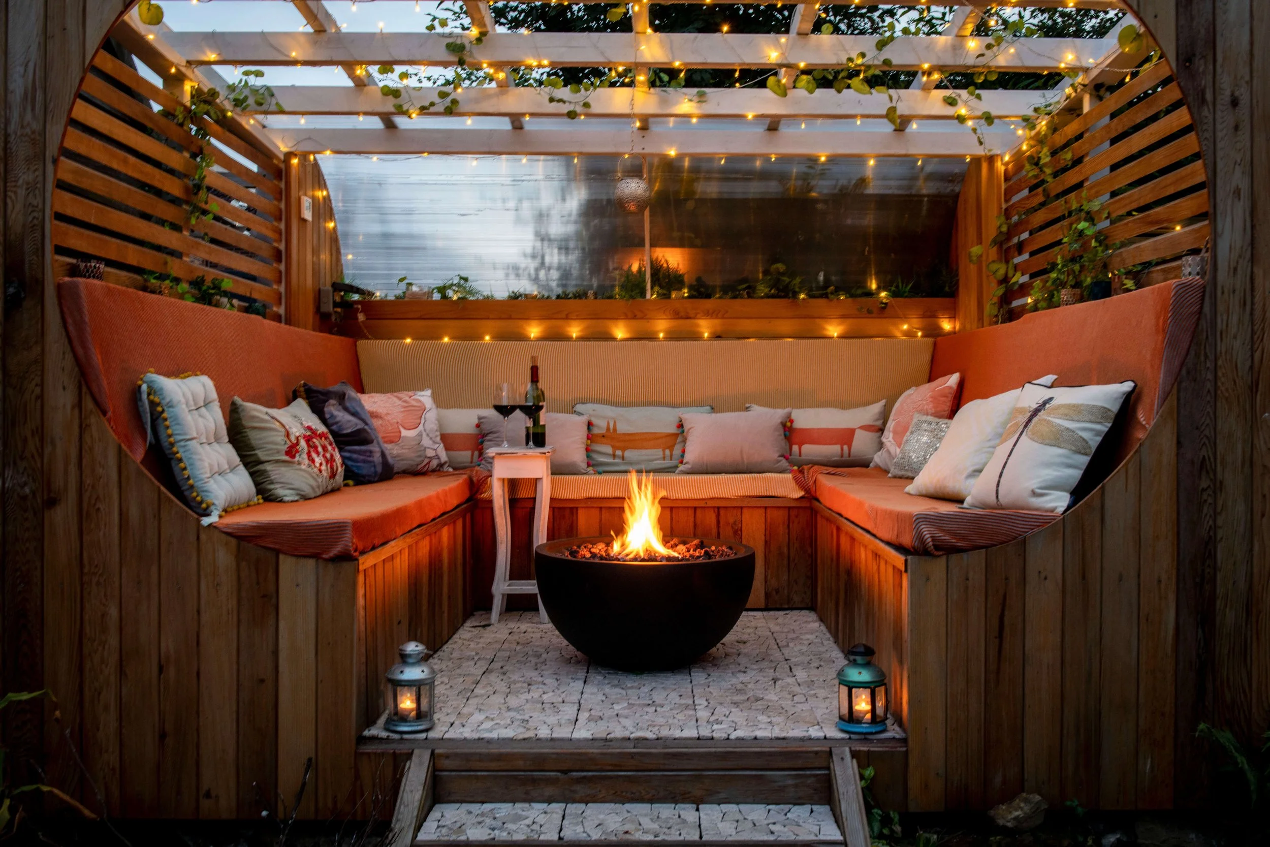 Cozy outdoor patio with built-in wooden bench seating, cushions, a small fire pit, string lights, and lanterns during evening.