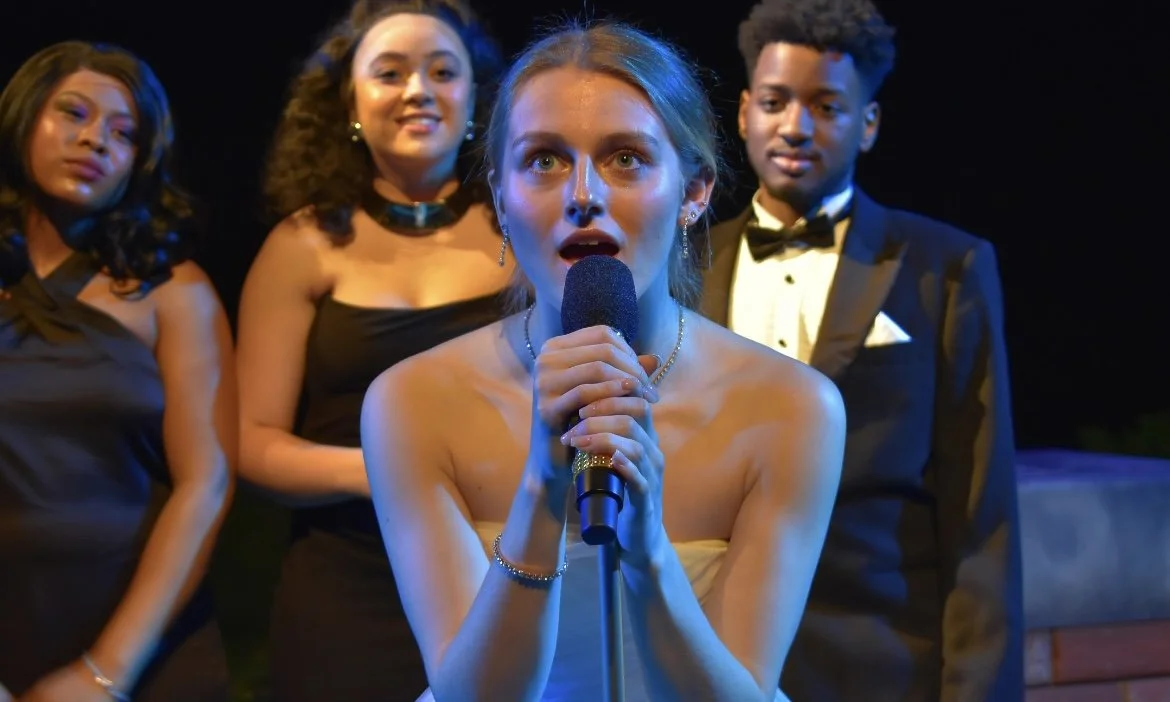 A woman in a strapless dress holding a microphone, with three people dressed formally behind her, on a stage with dark background.