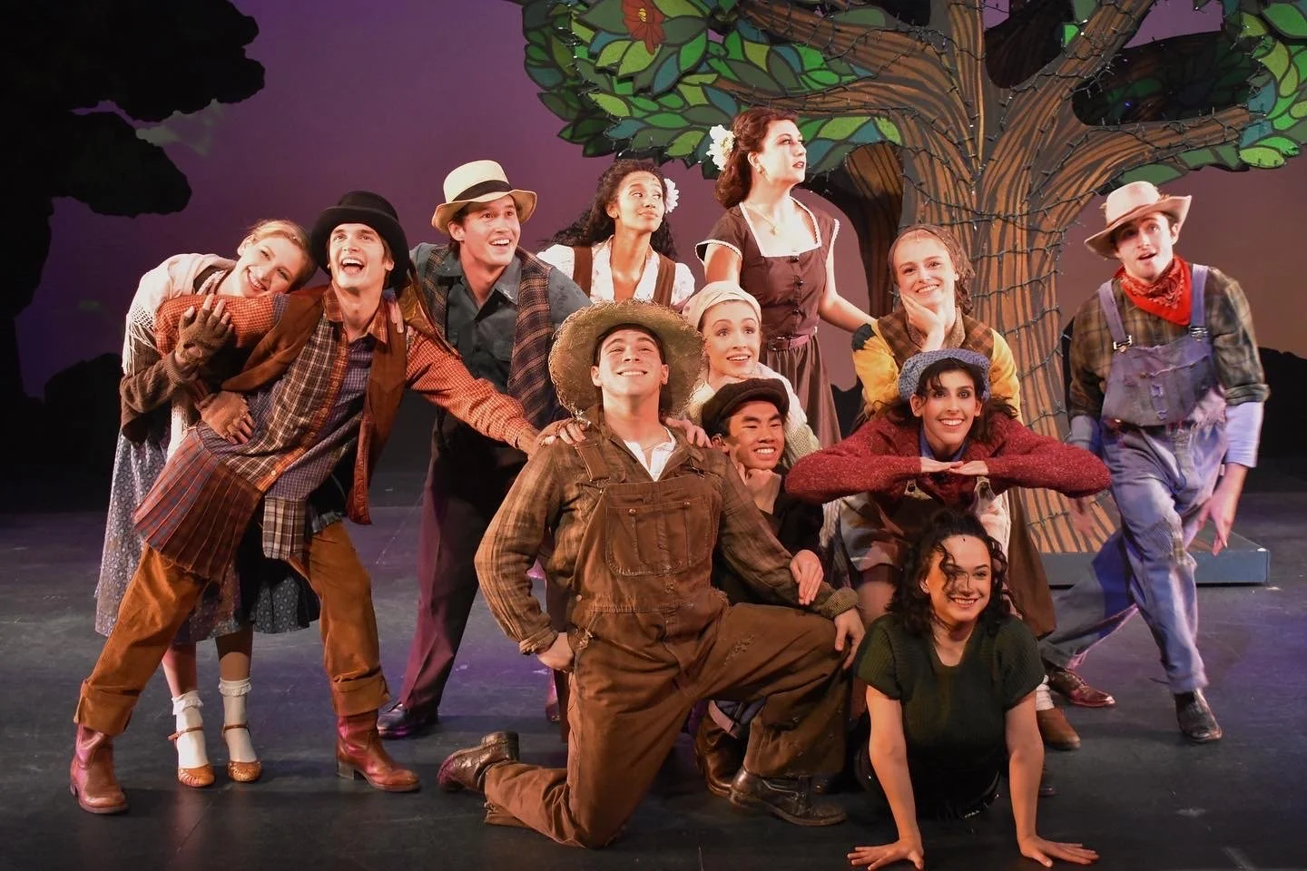 Group of actors in costume posing on stage in front of a painted backdrop of a tree, smiling and making playful gestures.