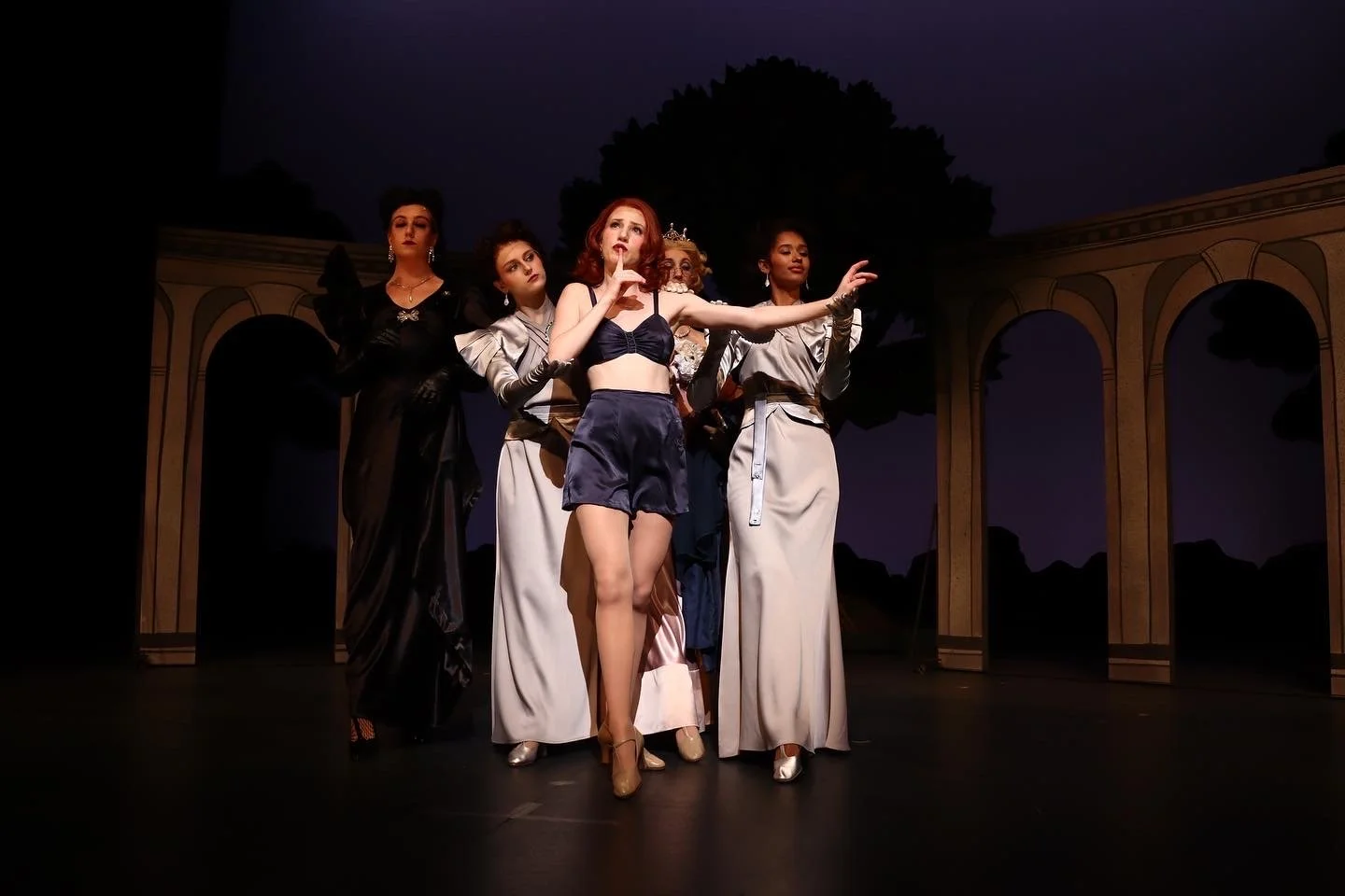 A scene from the musical 'As You Like It', standing on stage with the backdrop of an outdoor archway, elegant costumes, and dramatic lighting.