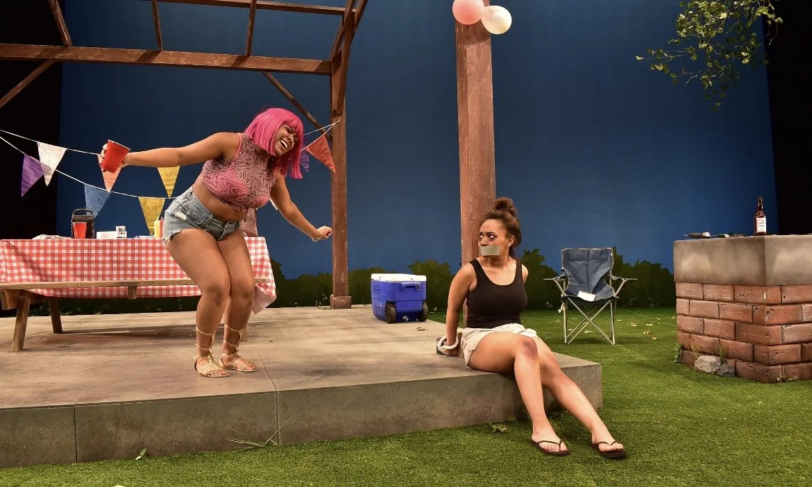 Two women at a party: one with pink hair jumping and the other seated with tape over her mouth.