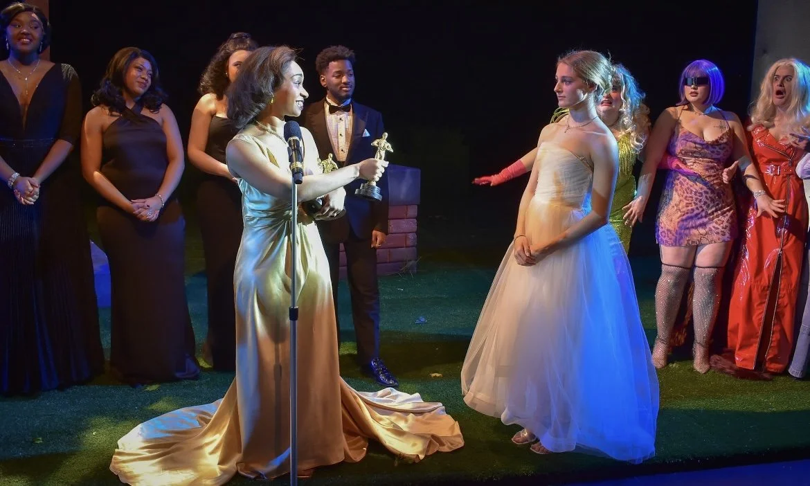 Women on stage during a ceremony, with one woman in a gold gown holding a trophy and speaking into a microphone, surrounded by other women dressed in evening wear and costume, some clapping and watching.