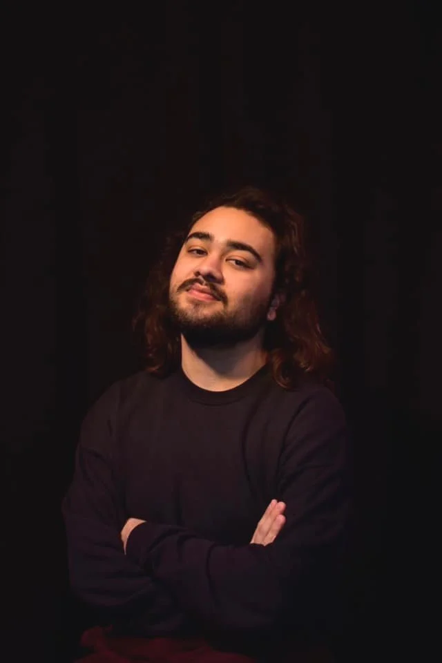 A young man with shoulder-length wavy hair, a beard, and a mustache, standing with arms crossed against a dark background, wearing a black long-sleeve shirt, looking confidently into the camera.