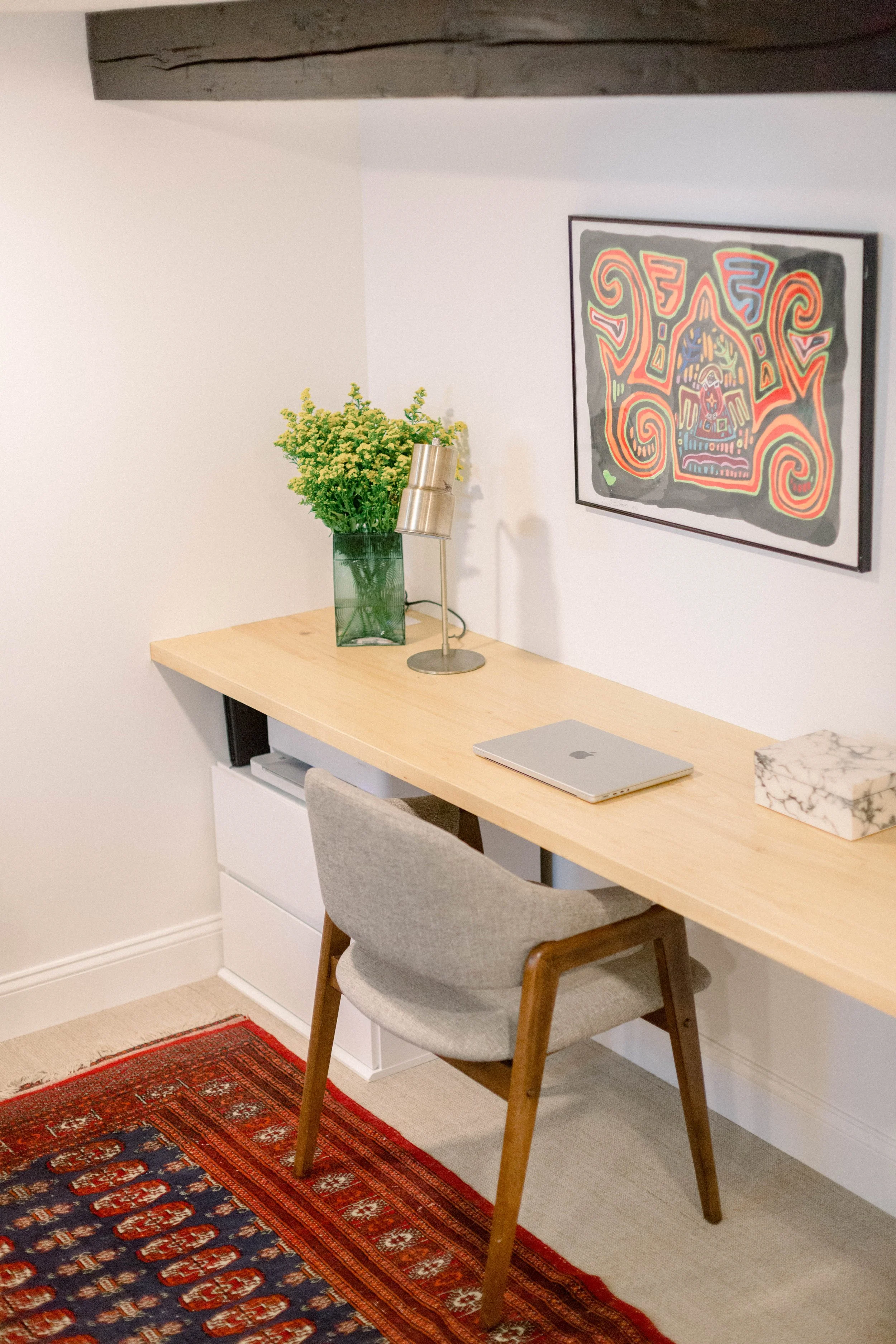 A home office desk area with a wooden desk, gray upholstered chair, a closed MacBook, a green vase with yellow flowers, a brass desk lamp, a marble tissue box, and a colorful abstract artwork on the wall.