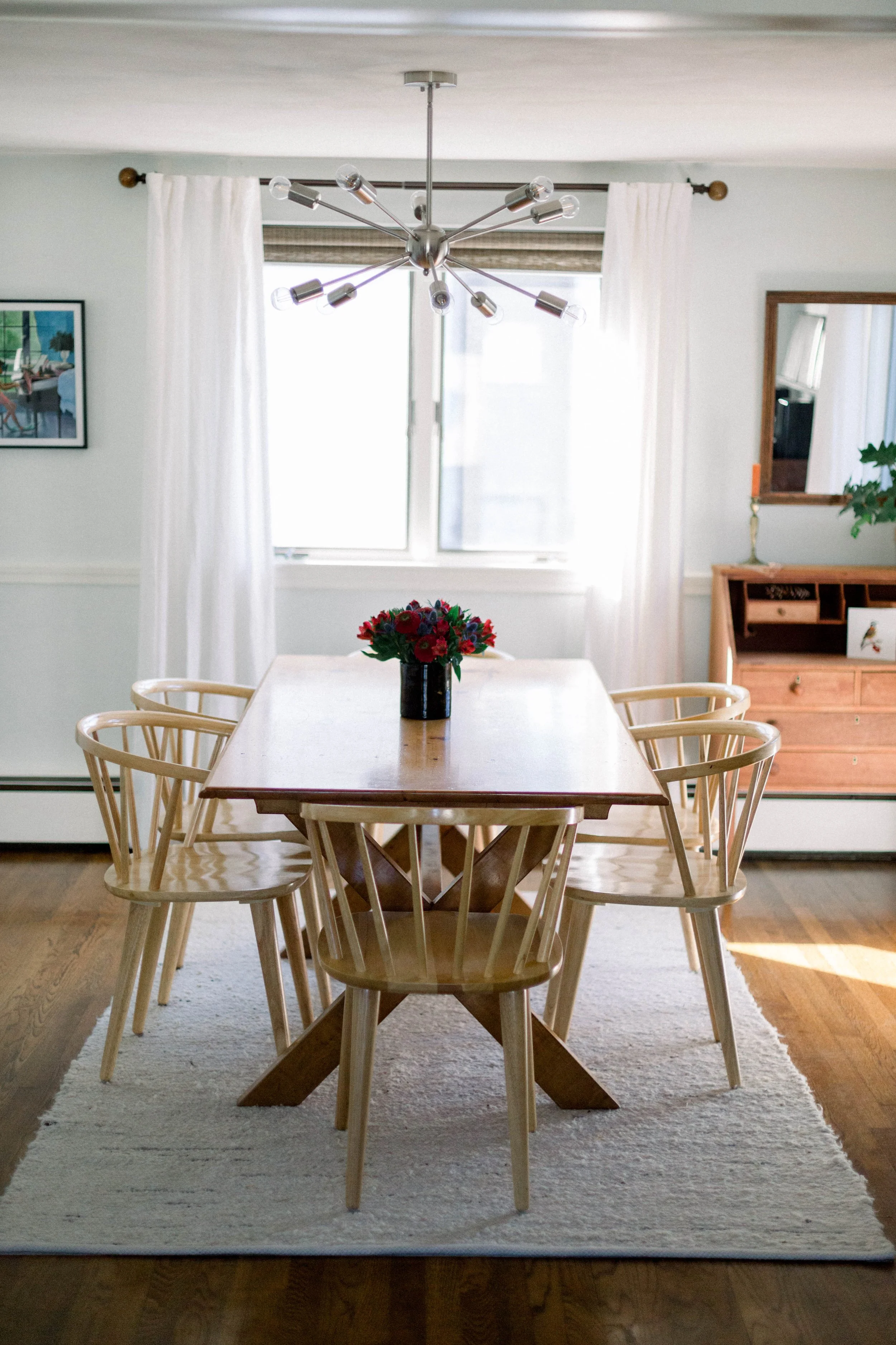 A dining room with a wooden table, six wooden chairs, a floral centerpiece, a window with white curtains, a modern chandelier, a small wooden sideboard, a mirror, and a framed picture on the wall.
