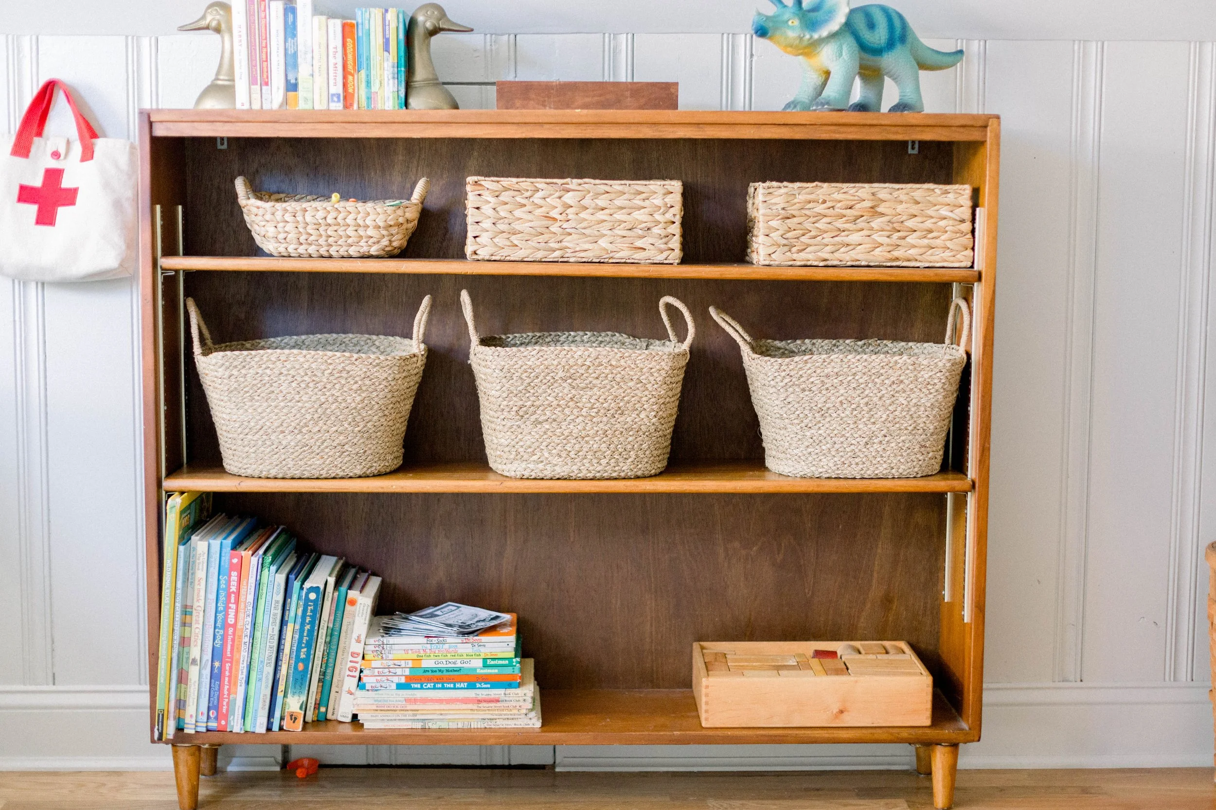 A wooden bookshelf with three shelves filled with baskets and books. The top shelf has a small basket and two woven dog-shaped figurines. The middle shelf has three large woven baskets. The bottom shelf has a stack of children's books and a wooden pu