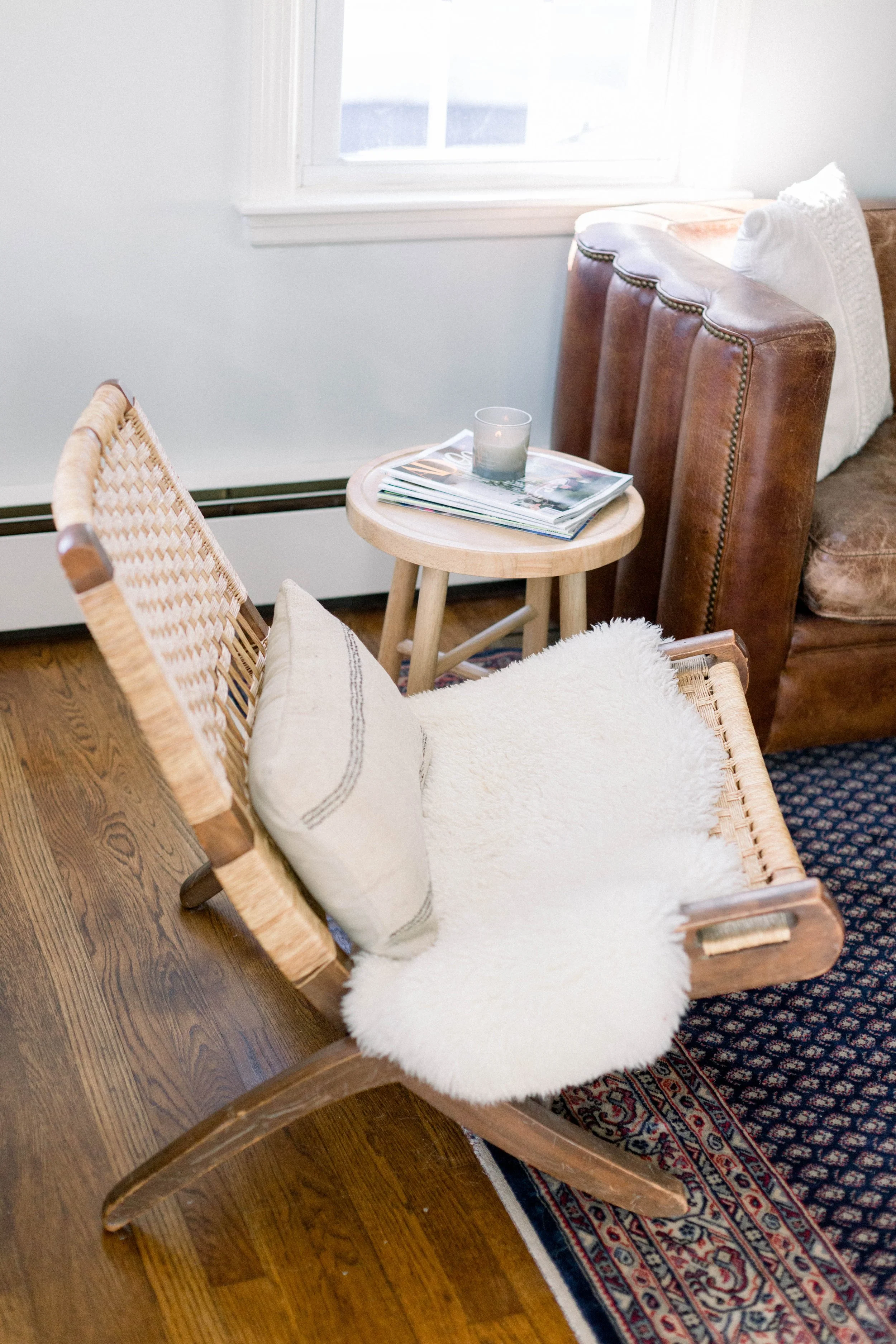 A cozy living room corner with a wooden chair draped with a white fluffy throw and a pillow, a round wood side table with magazines and a candle, and a leather couch with a white pillow, next to a window with white trim.
