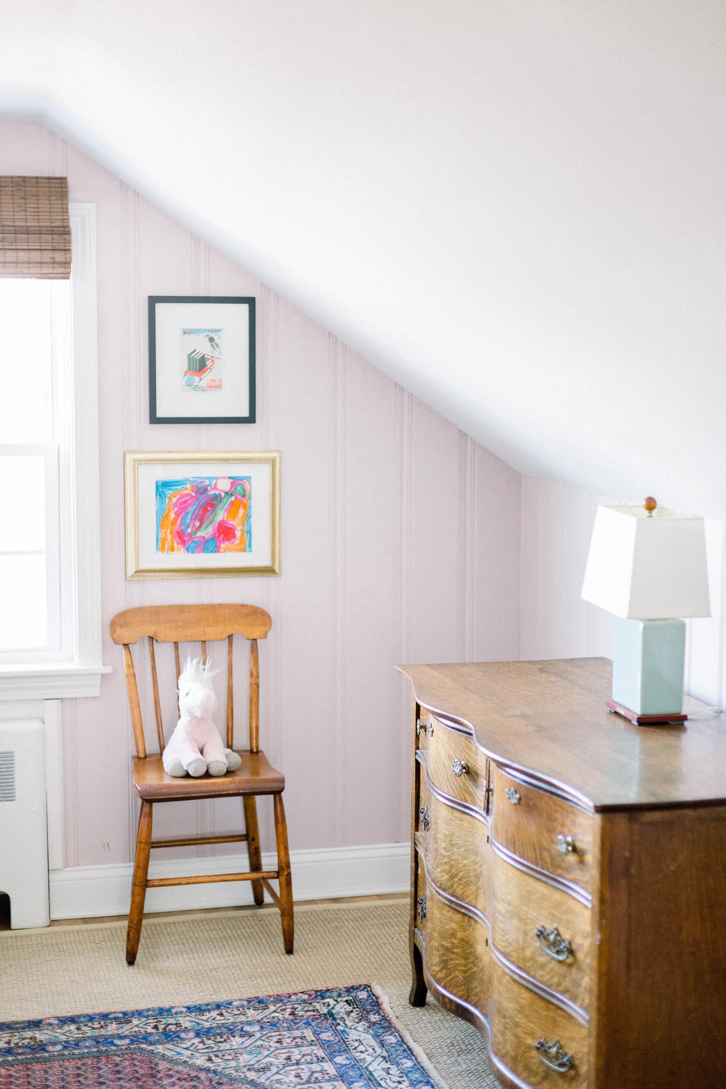 A room with a wooden chair, a plush unicorn toy on it, a window with a shade, framed artwork, a dresser, and a lamp. The wall has a sloped ceiling.