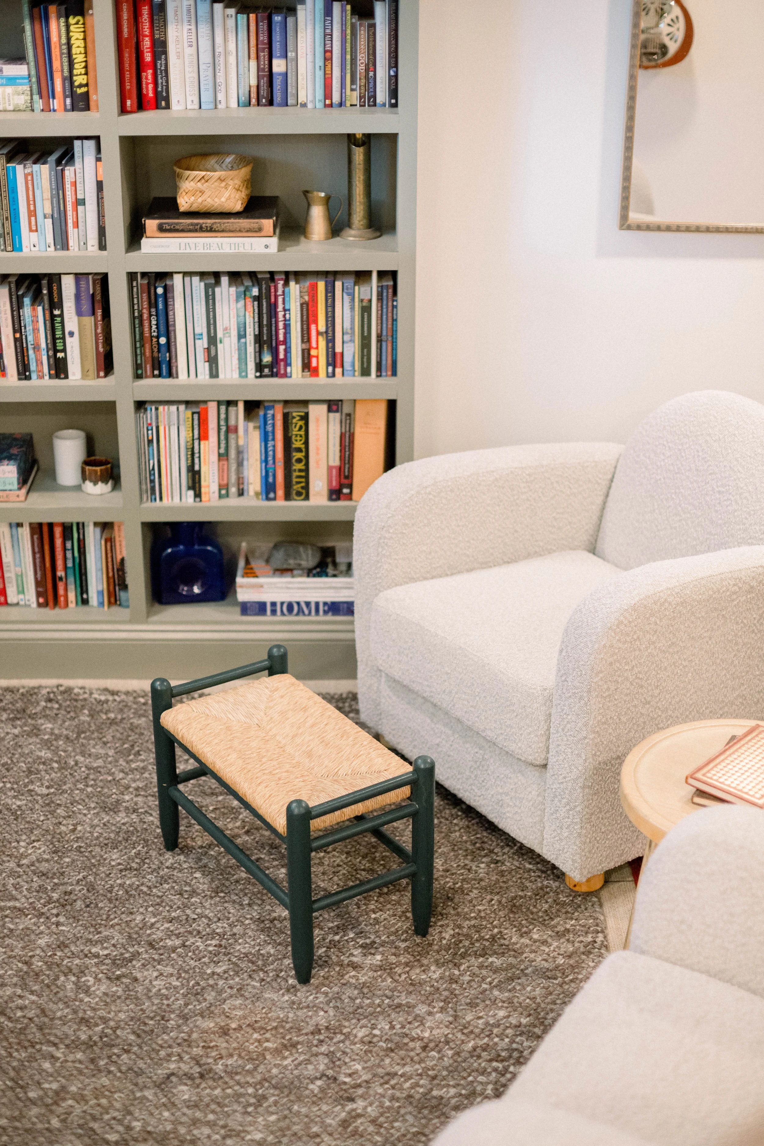 Living room with a beige armchair, a small black wooden footstool with a woven seat, a bookshelf filled with books, and a side table with a few items.