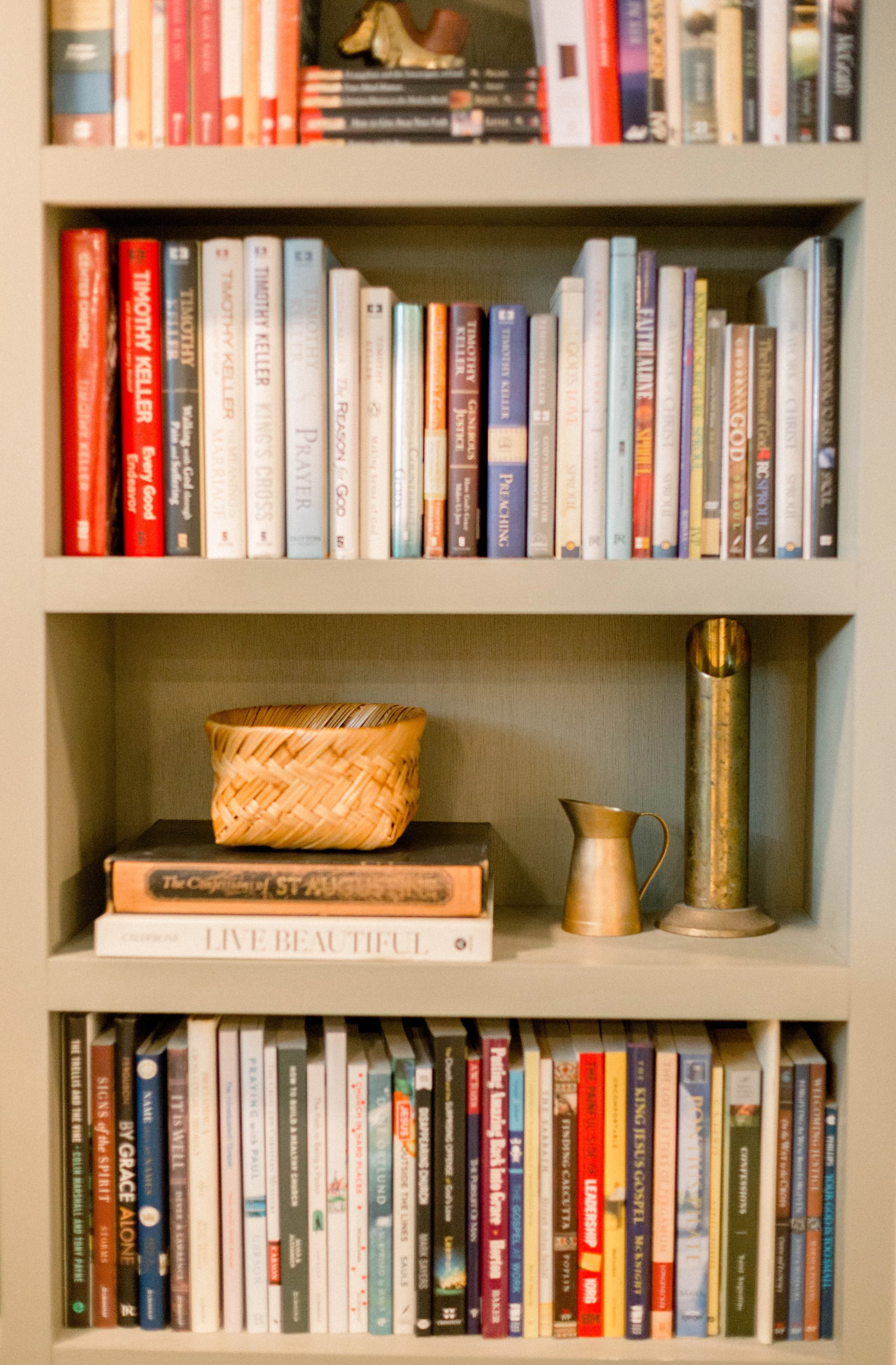 Bookshelf with various books, a woven basket, a gold-colored pitcher, a small gold-colored container, and a book titled 'Live Beautiful'.