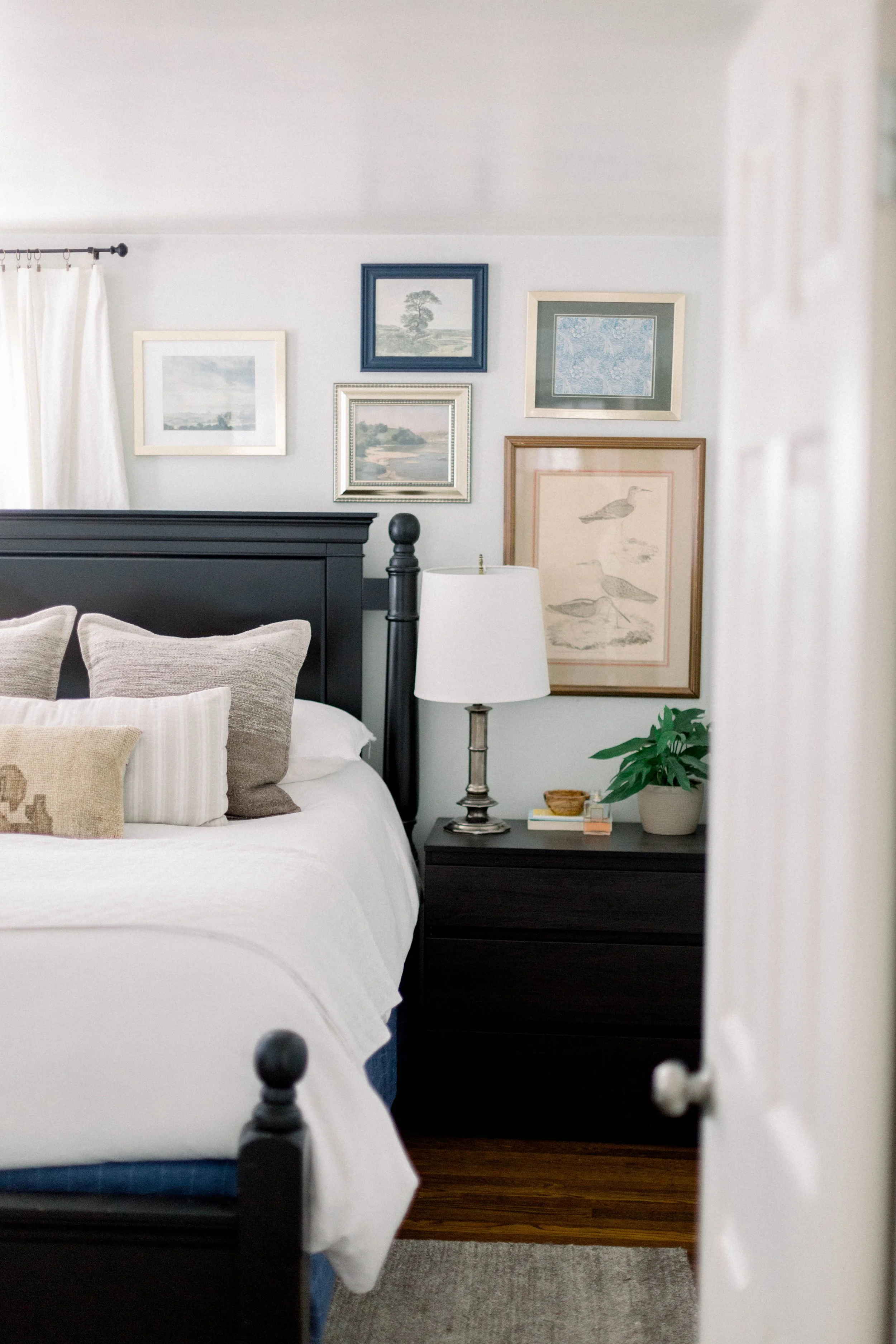 A bedroom with a black bed frame and white bedding, decorative pillows, a dark wood nightstand with a lamp, framed artwork on the light-colored wall, and a potted plant.