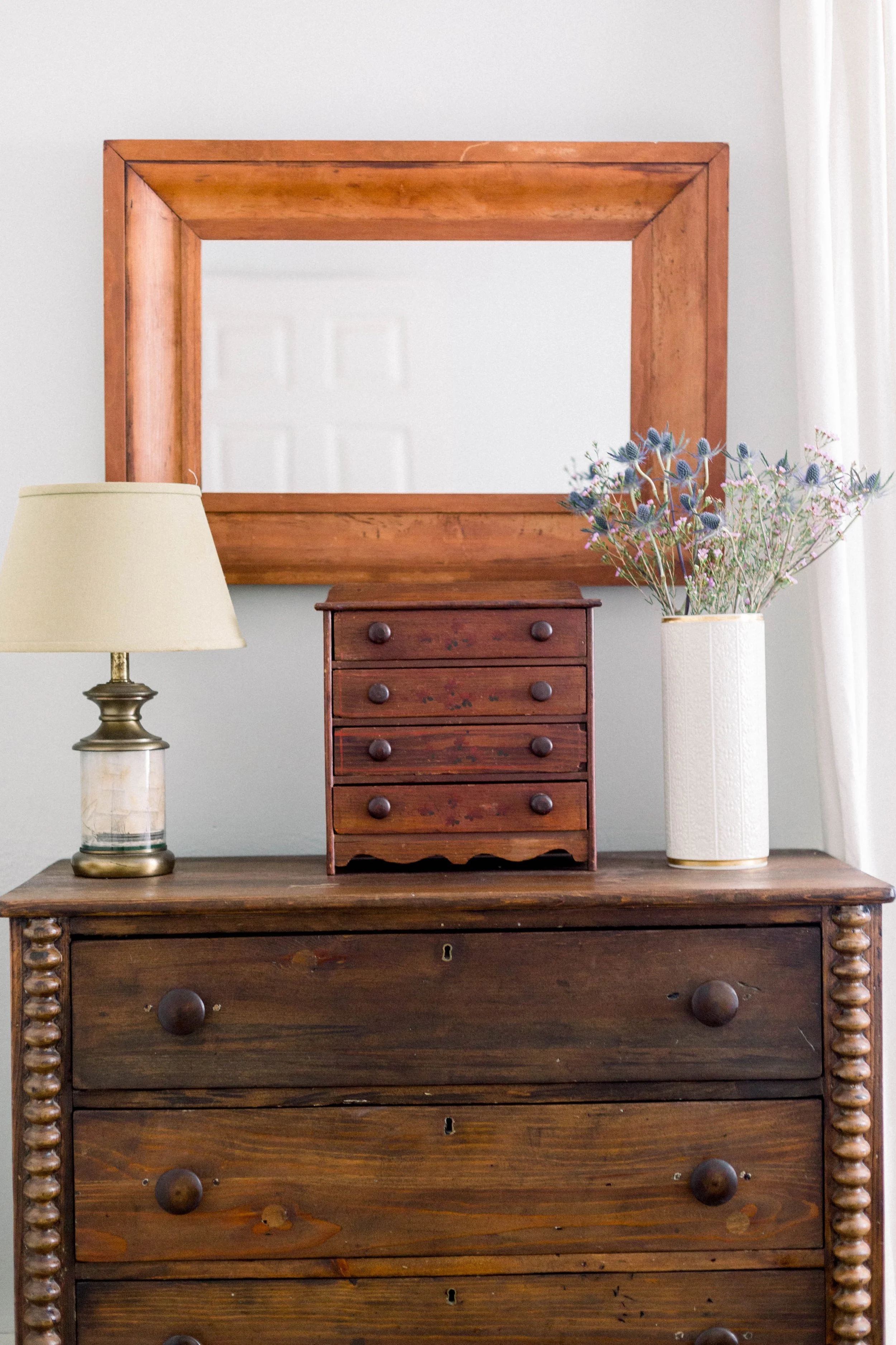 A wooden dresser with a mirror hanging above it. On the dresser, there is a table lamp with a beige lampshade on the left and a white vase with purple and green flowers on the right. A small wooden chest with five drawers is placed on top of the dres