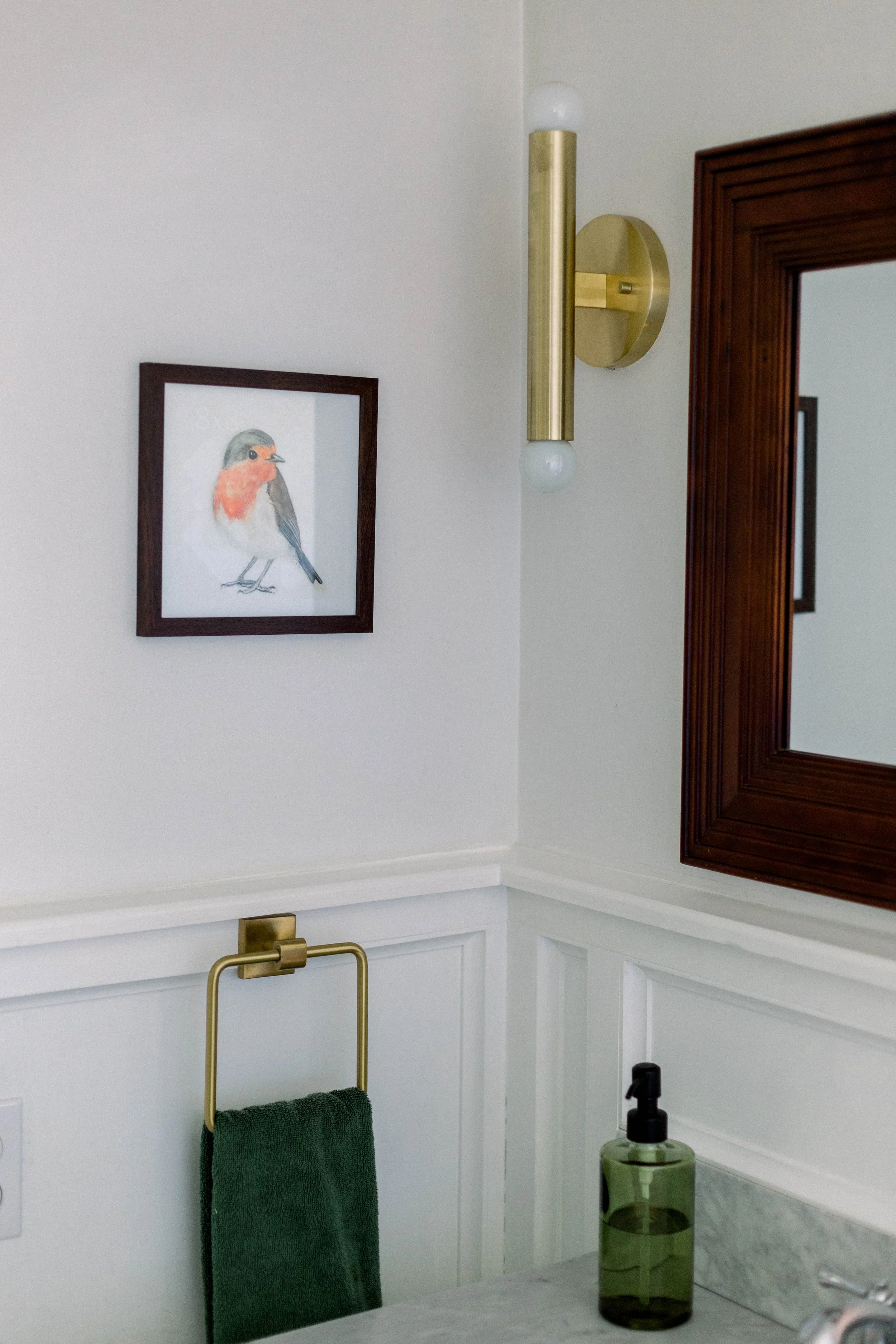 A bathroom corner with a framed bird photo on a white wall, a gold and white wall sconce, a green towel on a gold towel ring, a green soap dispenser, and a wooden framed mirror.