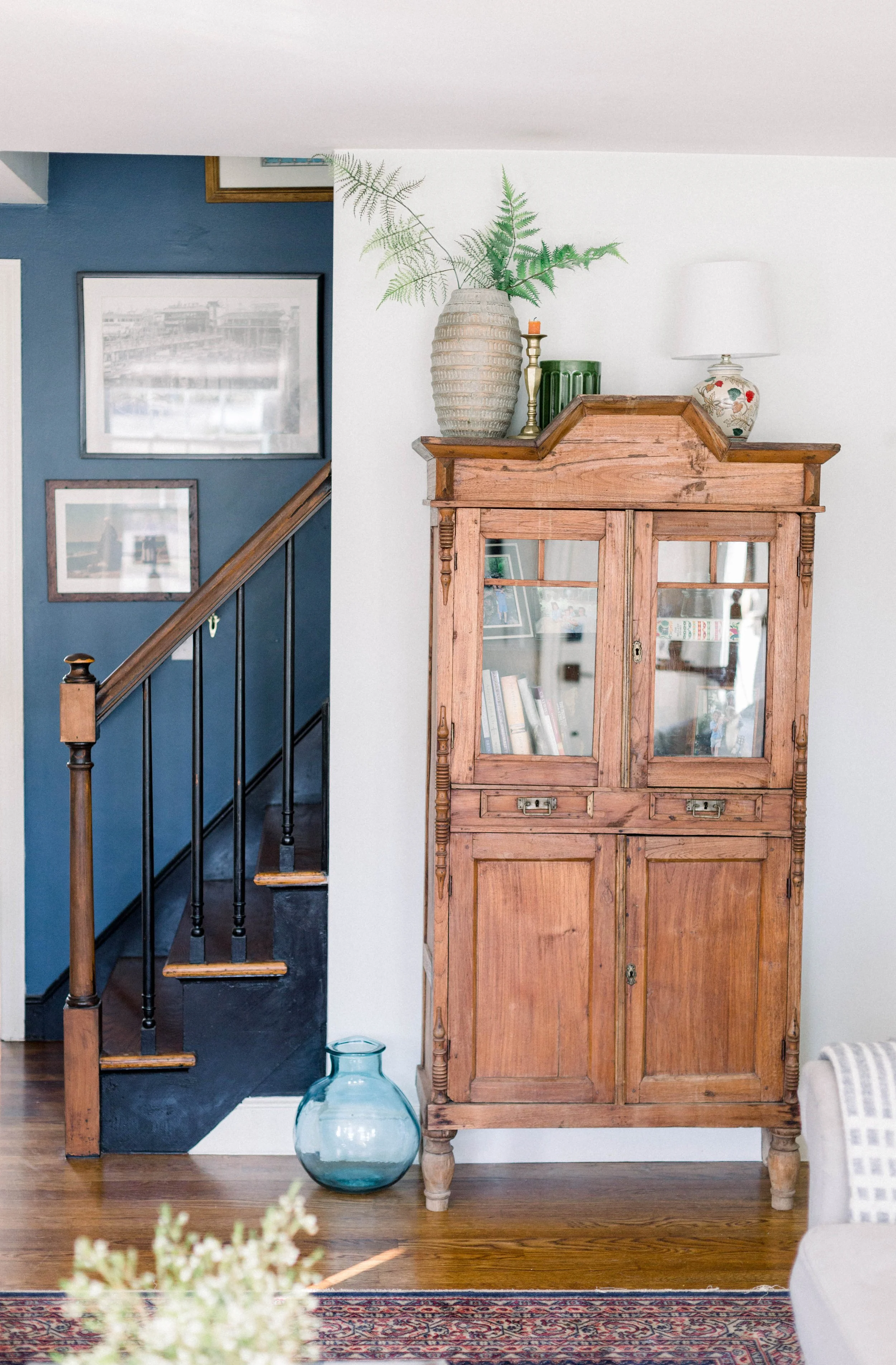 A wooden display cabinet with glass doors in a living room, decorated with a large vase of green fern leaves, a lamp, and other decorative items on top. A staircase with a blue wall and framed pictures is visible to the left, and there is a glass vas
