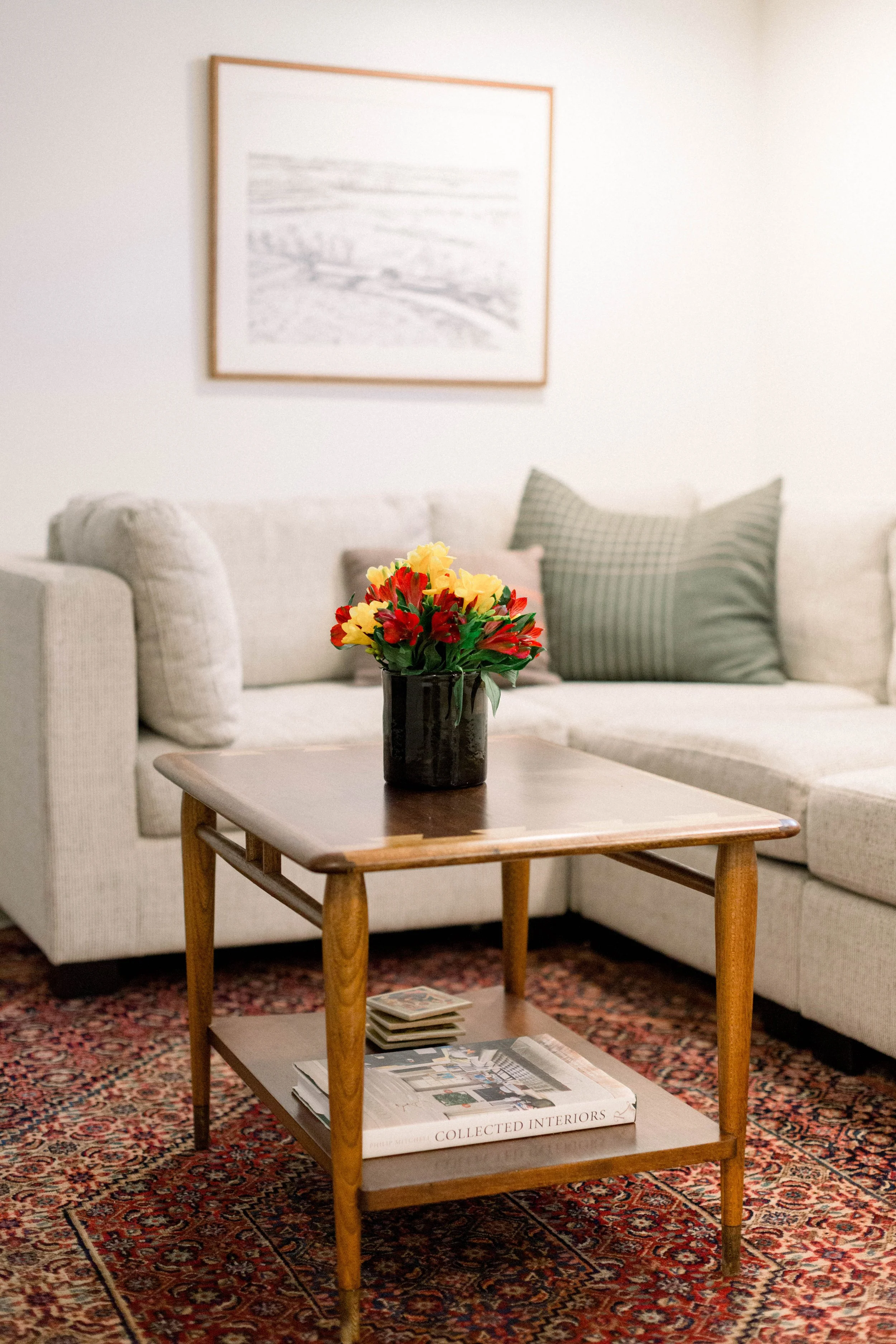 Living room with beige sofa, green plaid pillow, framed artwork, wooden coffee table with a black vase of red and yellow flowers, and a patterned rug.