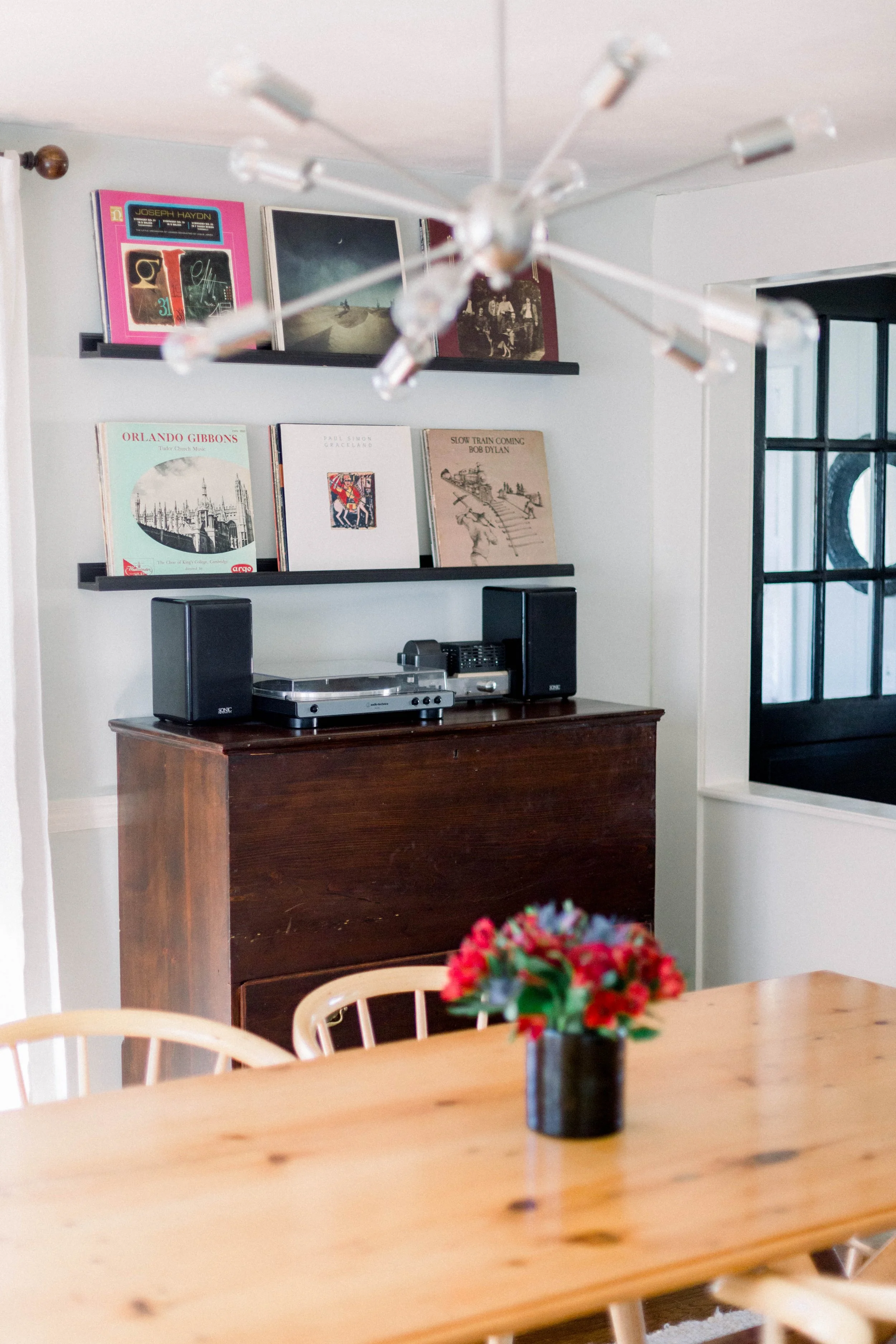 View of a dining area with a wooden table and a small potted flower, a stereo system on a wooden sideboard, and two black wall-mounted shelves with vinyl records and framed artwork on the white wall.