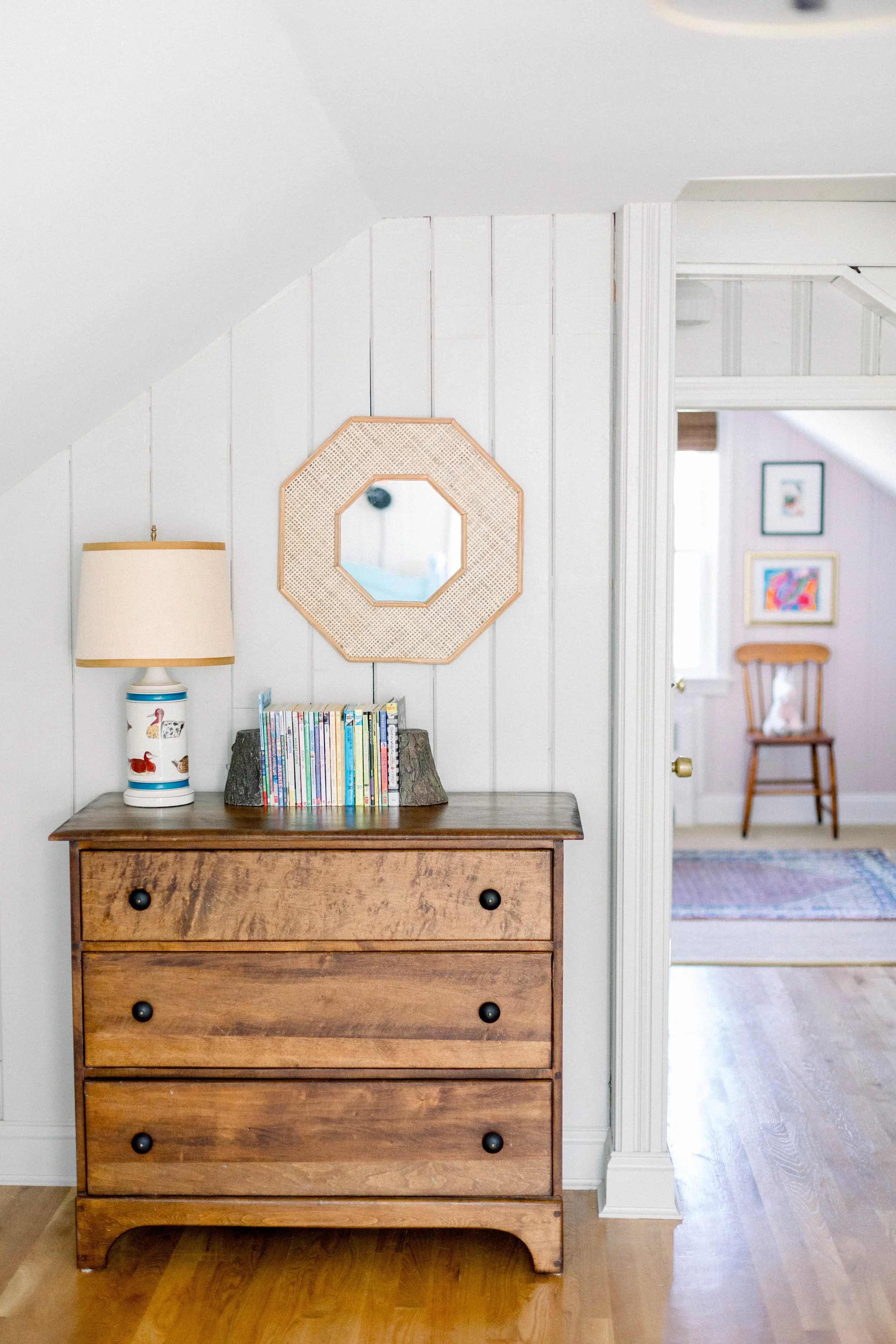 A wooden dresser with a lamp and a row of children's books on top, beside a mirror on the wall.