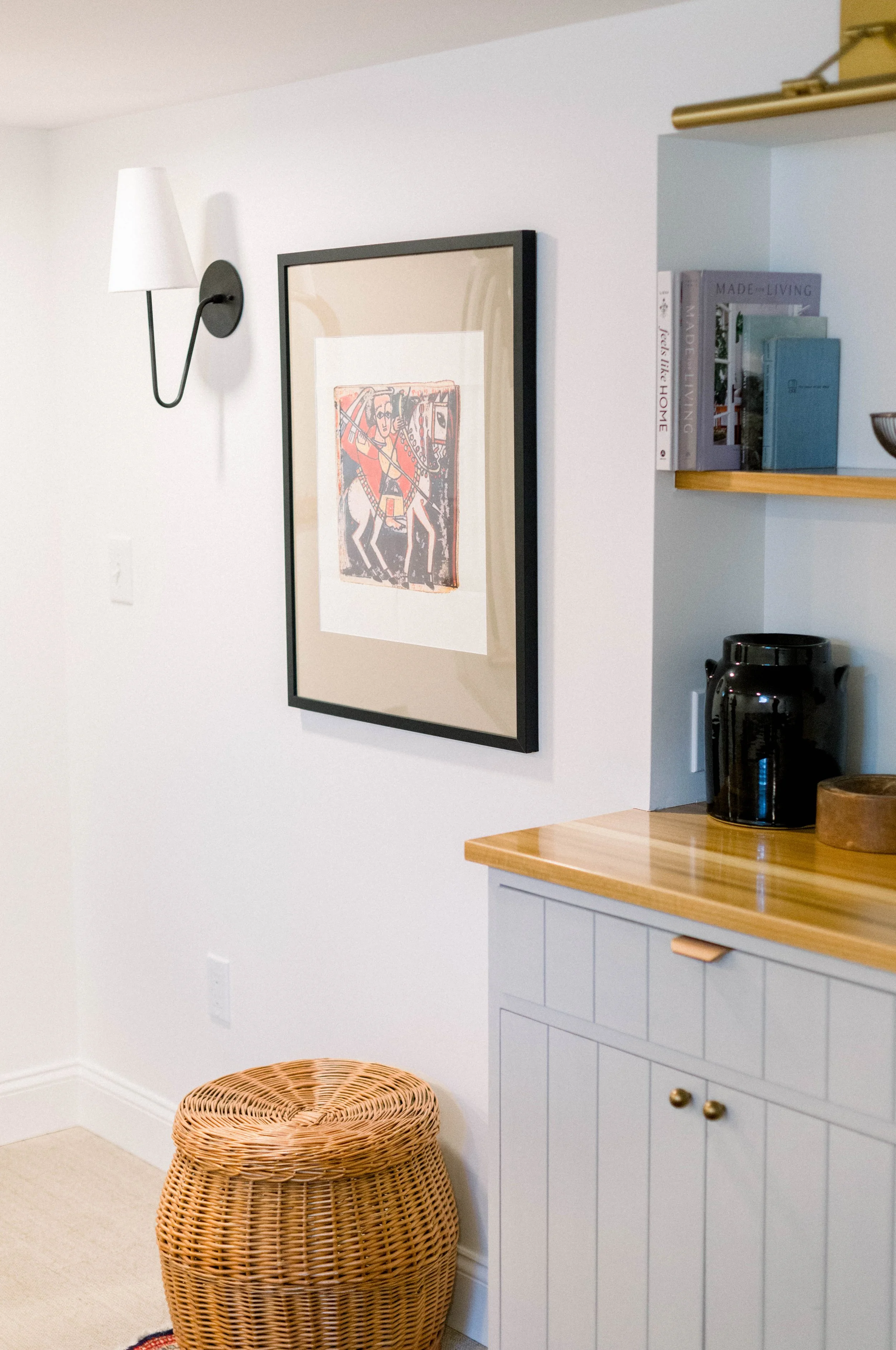 A framed artwork on a white wall, a wall-mounted lamp, a wicker ottoman, and part of a kitchen cabinet with black appliances and books.