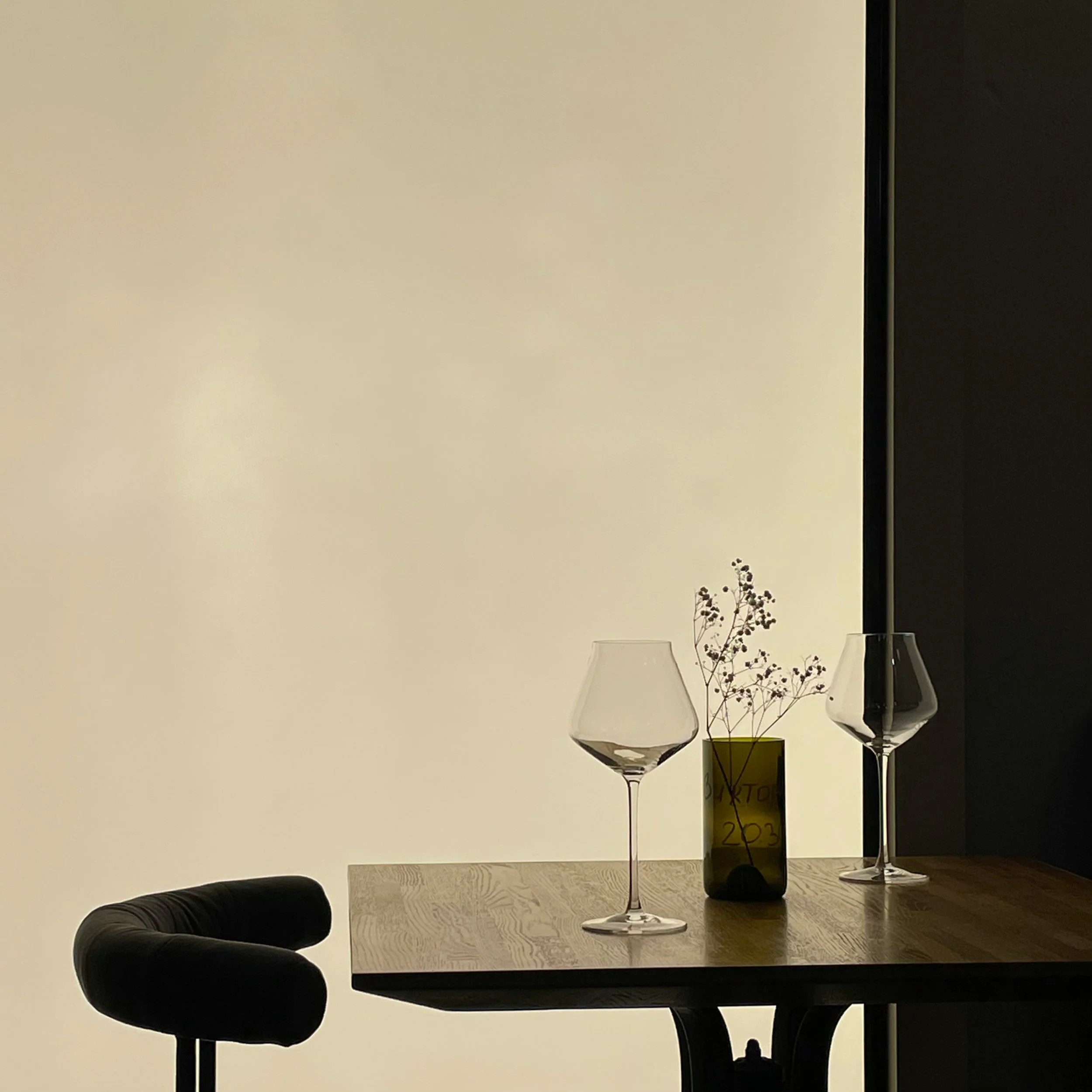 A wooden table with two empty wine glasses and a small vase with dried flowers, set against a minimalistic background with a cream-colored wall and dark shadowed area.