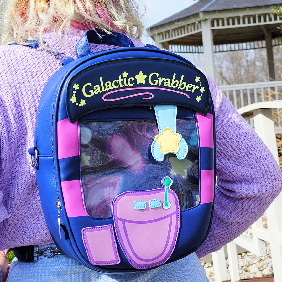Child's backpack with a space-themed design, featuring the text 'Galactic Grabber' and colorful patches, including a star-shaped zipper pull, worn by a person in a purple sweater outdoors.